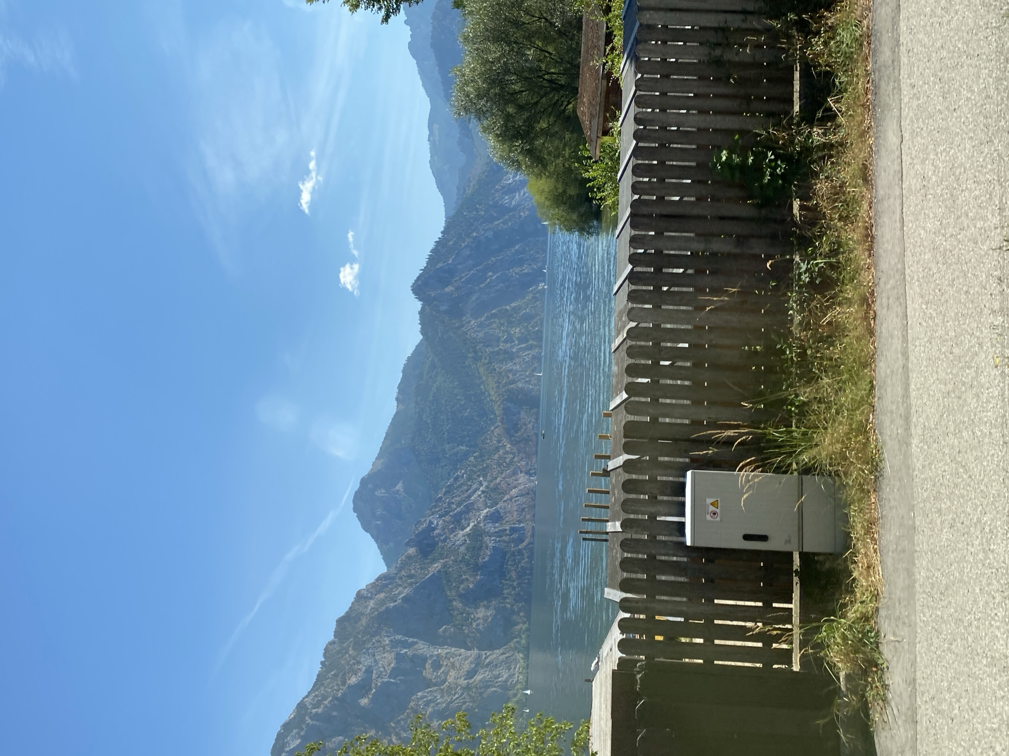 A wide shot captures a serene lake scene with majestic mountains in the background under a clear blue sky. In the foreground, a weathered wooden fence runs horizontally across the frame, partially obscuring a light-colored electrical box. Beyond the fence, the calm, reflective surface of a lake is visible, dotted with a few sailboats. The lake is embraced by steep, rocky mountains covered with sparse vegetation, their peaks stretching towards the sky. On the right side of the image, lush green trees and a small wooden structure with a brown roof are nestled near the shoreline. The overall atmosphere is tranquil and picturesque, suggesting a peaceful day in a natural setting. This scene is located in Altmünster, Austria.