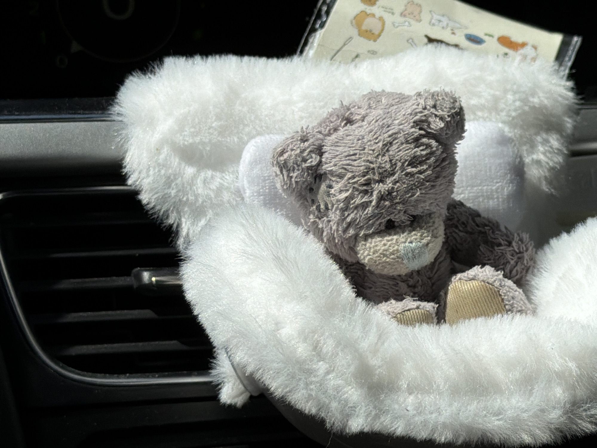 A stuffed grey teddy bear sits nestled within a white, fluffy steering wheel cover inside of a vehicle. The steering wheel itself is mounted on the dashboard, positioned in front of an air vent. Above the bear, a piece of paper with illustrations is visible. The location and city cannot be determined from the image.