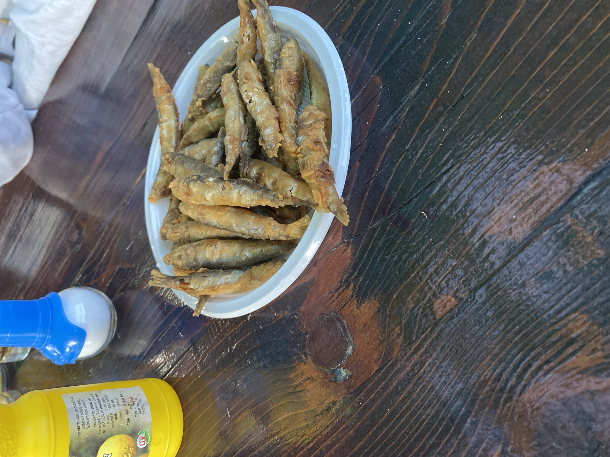 A close-up, overhead shot shows a white paper plate filled with small, golden-brown fried fish resting on a dark wooden table. The fish are whole and appear to be a type of smelt or sardine, lightly battered and fried to a crispy texture. Some salt granules are visible on the table surface. To the left of the plate is a yellow plastic squeeze bottle, likely containing a sauce or condiment, with text in Cyrillic. Next to the bottle is a clear glass salt shaker with a blue cap. The table is made of rough-hewn, dark wood planks with visible grain and knots. In the background, out of focus, is a white cloth and a hint of what might be a glass. The setting appears to be outdoors, possibly a casual dining area. While the image does not explicitly show Smolyan, Bulgaria, the presence of Cyrillic text on the bottle suggests a location where this alphabet is used, such as Bulgaria.
