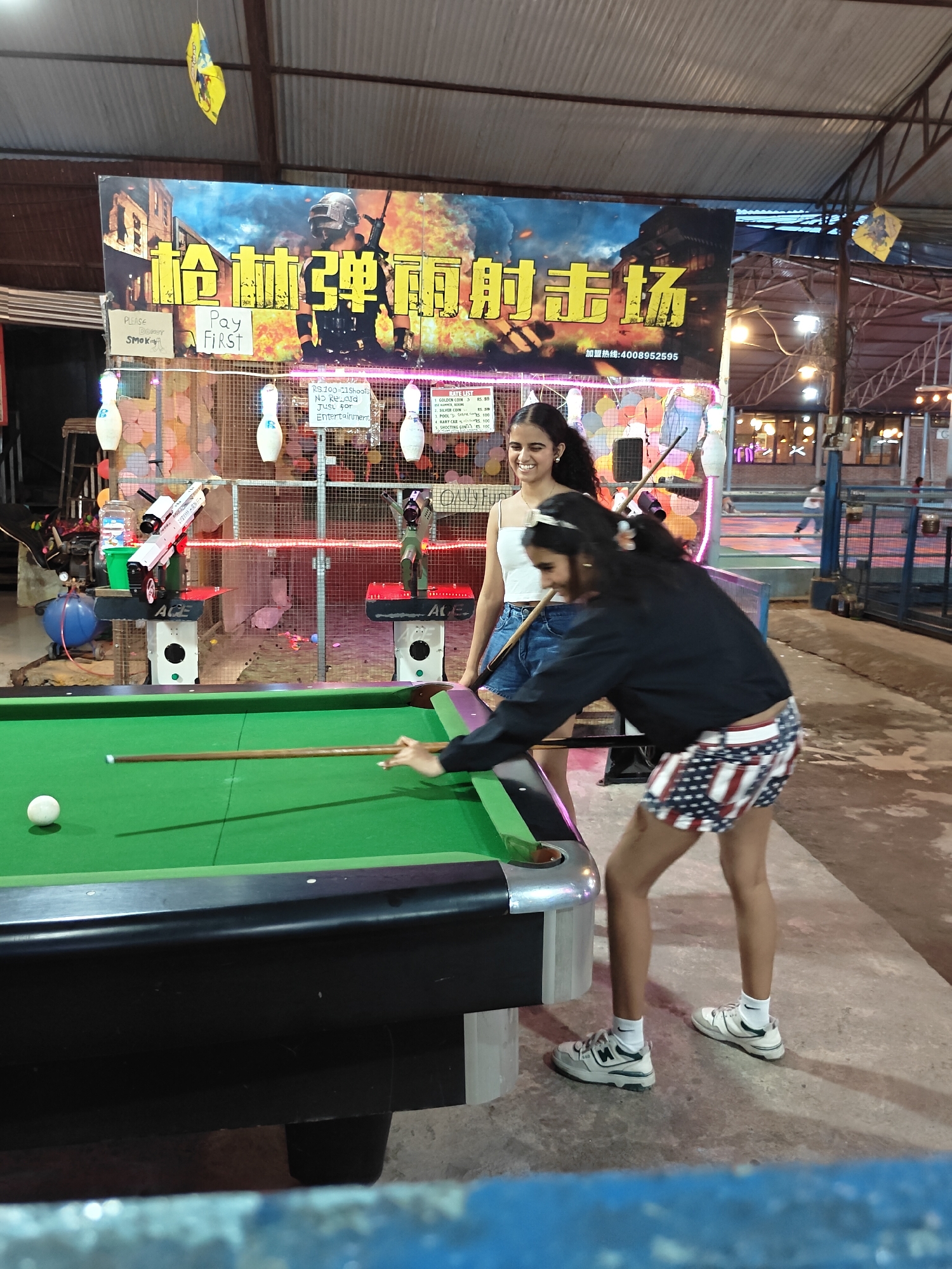 In Pokhara, Nepal, two young women are playing pool in an outdoor amusement area. The woman on the left is wearing a white tank top and denim shorts, while the woman on the right is wearing a black jacket and patriotic-themed shorts. A pool table with green felt and a single white ball is in the foreground. Behind them, a banner with Chinese characters and imagery of a soldier is displayed. The area appears to be an entertainment venue, with game stalls and neon lights visible in the background.