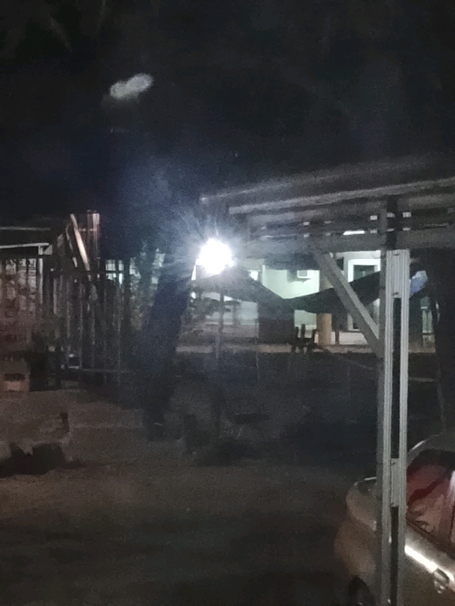 The image shows a dimly lit outdoor scene at night, possibly in Ujungrusi, Indonesia. In the foreground, a portion of a silver car is visible on the right, with a person wearing a red garment visible inside. A metal structure, likely part of a carport or awning, extends across the upper portion of the frame. In the mid-ground, a bright light source illuminates a dark tree trunk and casts shadows. Behind the tree, a building with windows and an air conditioning unit is partially visible. Fencing or a similar structure is present to the left of the tree. The ground appears to be paved or a dirt surface. A faint, amorphous light anomaly is present in the dark sky area in the upper left of the image. The overall impression is of an urban or semi-urban environment at night, with artificial lighting being a dominant feature.