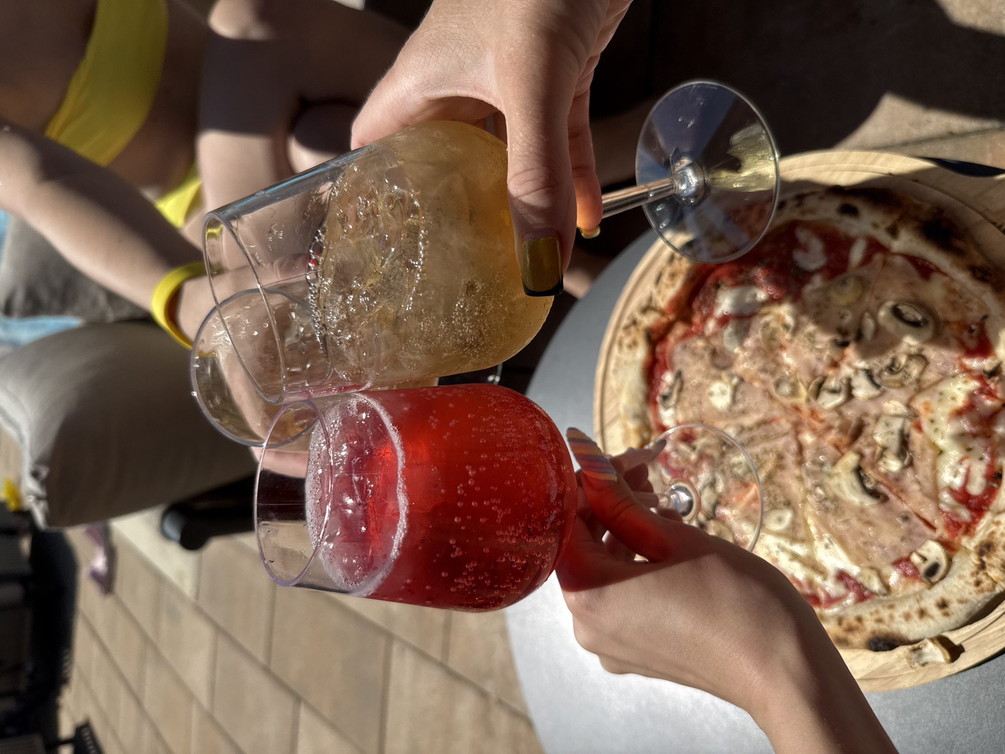 An outdoor, sunlit scene depicts three individuals engaging in a toast. Three hands are shown holding stemmed glasses, clinking them together above a pizza. The hand on the left is pale with long, multi-colored (rainbow-striped) fingernails, gripping a glass containing a vibrant red, carbonated beverage with ice cubes. The hand on the right is tanned with yellow fingernails featuring black tips, holding a glass filled with a light amber, carbonated beverage with ice cubes. A third, partially obscured hand holds a similar clear glass in the center.
Below the clinking glasses, a large, round, freshly cooked pizza is presented on a wooden serving board. The pizza features a thin, slightly charred crust and is topped with tomato sauce, melted white cheese, sliced mushrooms, and what appears to be thinly sliced ham or prosciutto. The pizza and serving board rest on a dark gray, circular table.
The background elements include light-colored paver stones or tiles on the ground, a beige outdoor furniture cushion, and a section of blue water, suggesting a swimming pool. Strong, defined shadows are cast across the scene, indicating bright, direct sunlight. In the upper right background, a person wearing a yellow bikini top and a yellow wristband is partially visible. Further in the distant background to the left, a pair of pink flip-flops is visible on the ground. The specific location within the city of Unknown, Unknown, is an outdoor leisure area, likely a patio or poolside deck.