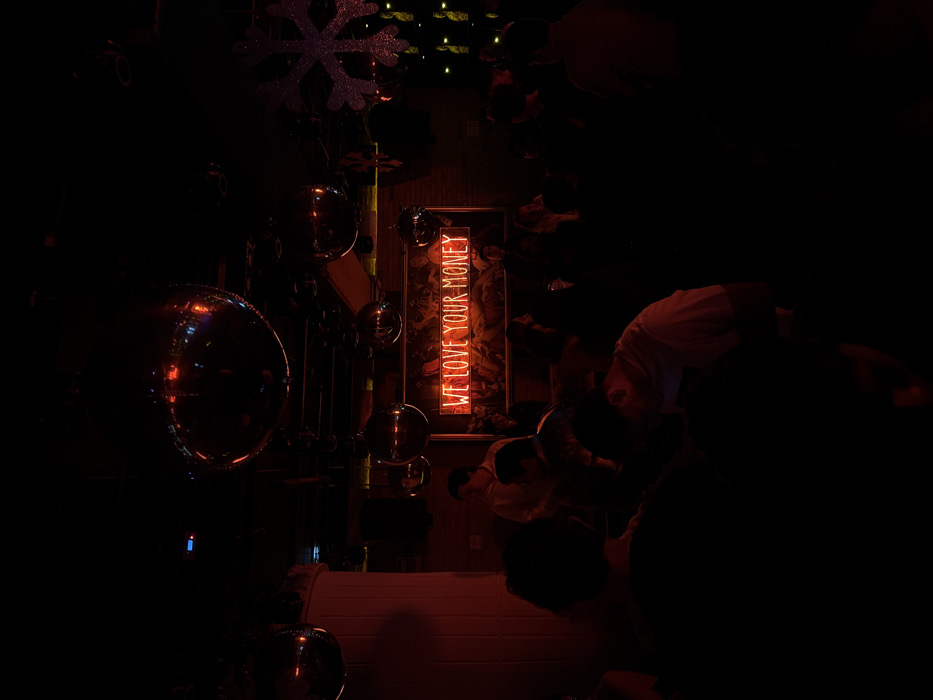 The image depicts an interior scene, likely a bar or nightclub, at night, characterized by deep red ambient lighting. The setting is San Bernardino Tlaxcalancingo, Mexico. The ceiling features multiple large, reflective disco balls, casting faint glints of light. A prominent red neon sign is mounted on a brick wall at the back, displaying the phrase "WE LOVE YOUR MONEY" in all capital letters. Below and around this sign, a large, dark framed artwork or mural is partially visible. To the far right, a glittery, white snowflake decoration is suspended from the ceiling.
Numerous individuals are present within the space, mostly appearing in dark silhouettes due to the low light conditions. They are standing and clustered throughout the room, facing various directions, suggesting a social gathering or event. Visible subjects include a man in a light-colored shirt standing near the center-left of the frame, with his back partially turned to the viewer. Other individuals are less distinct, visible as dark forms against the red-tinged background, with minimal discernible facial features or specific actions beyond standing and interacting. The overall environment suggests a lively, crowded atmosphere.