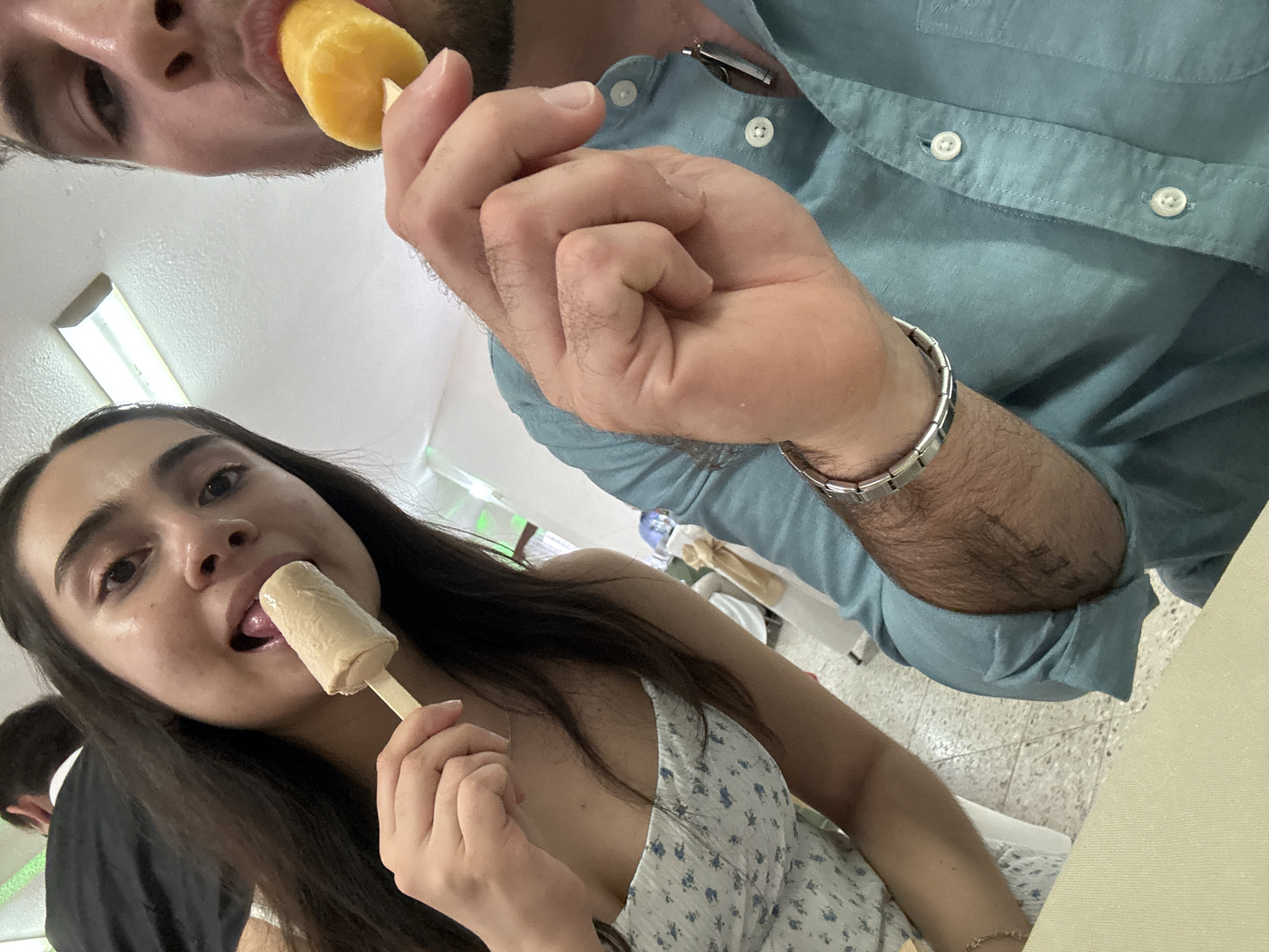 Two people are eating popsicles in what appears to be an indoor setting. A woman in the foreground is holding and licking a beige-colored popsicle. She has long, dark hair and is wearing a floral-patterned top. A man is partially visible in the background, also eating a popsicle. He's wearing a blue button-down shirt and a silver bracelet. The lighting is bright and even, suggesting artificial illumination. The background is out of focus, showing tables and chairs, and there's what appears to be a window with green and white light shining through. The setting seems to be a casual eatery or similar space. I cannot confirm the location in Chipilo de Francisco Javier Mina (Chipilo), Mexico with certainty based on the image alone.