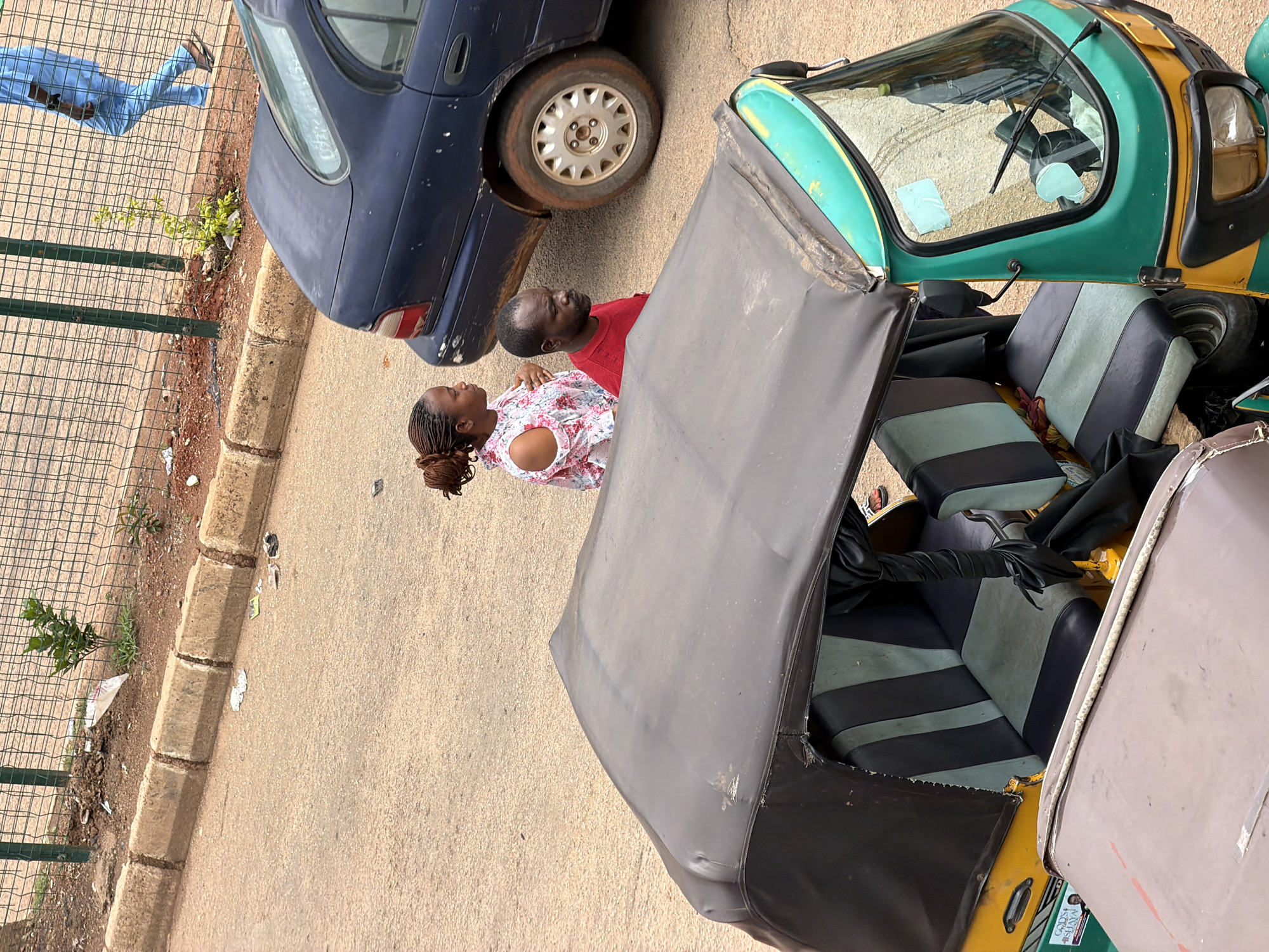 The image, taken from an elevated perspective in Bukuru, Nigeria, shows a street scene with two individuals standing near a dark blue car. The woman, with braided hair, wears a floral-patterned off-the-shoulder top, while the man is dressed in a red t-shirt. They are positioned to the left of the car, with the woman facing forward and the man looking to the side. In the foreground, the top of a three-wheeled auto-rickshaw is visible, featuring a black canopy and green trim. The street surface is made of asphalt, and a concrete curb runs along the edge of a dirt area with a wire fence behind it. Scattered debris is present on the ground near the curb. A person in a light blue outfit is seen walking along the sidewalk behind the fence.