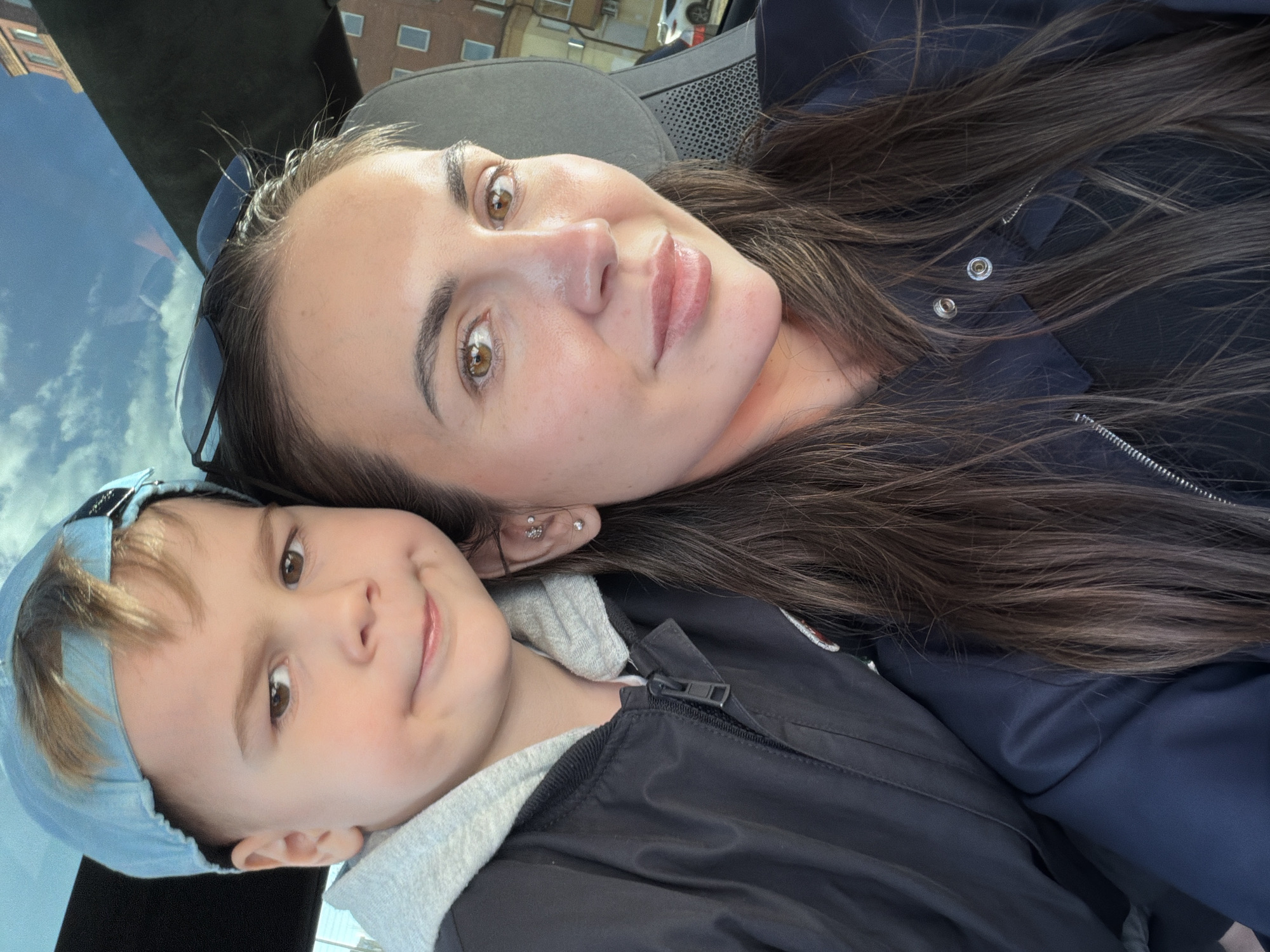 A close-up selfie captures a woman and a young child lying down, seemingly in a car, with the image oriented sideways. The woman has long, dark brown hair, brown eyes, and wears a dark blue jacket. Sunglasses rest on top of her head, and she has several ear piercings, including a decorative stud. She looks towards the camera with a soft expression.
Beside her, a young boy with light brown hair wears a light blue baseball cap and a black zippered jacket with a grey hood. He also gazes towards the camera with a gentle, slightly smiling face. Their proximity suggests a close, familial relationship.
Reflections in a window or sunroof reveal a clear blue sky with white clouds and distant buildings, indicating an outdoor setting during daylight hours. A dark car seat and headrest are partially visible in the background. The overall atmosphere conveyed is calm and affectionate. The scene is set in Krasnoyarsk, Russia. There is no visible text in the image.