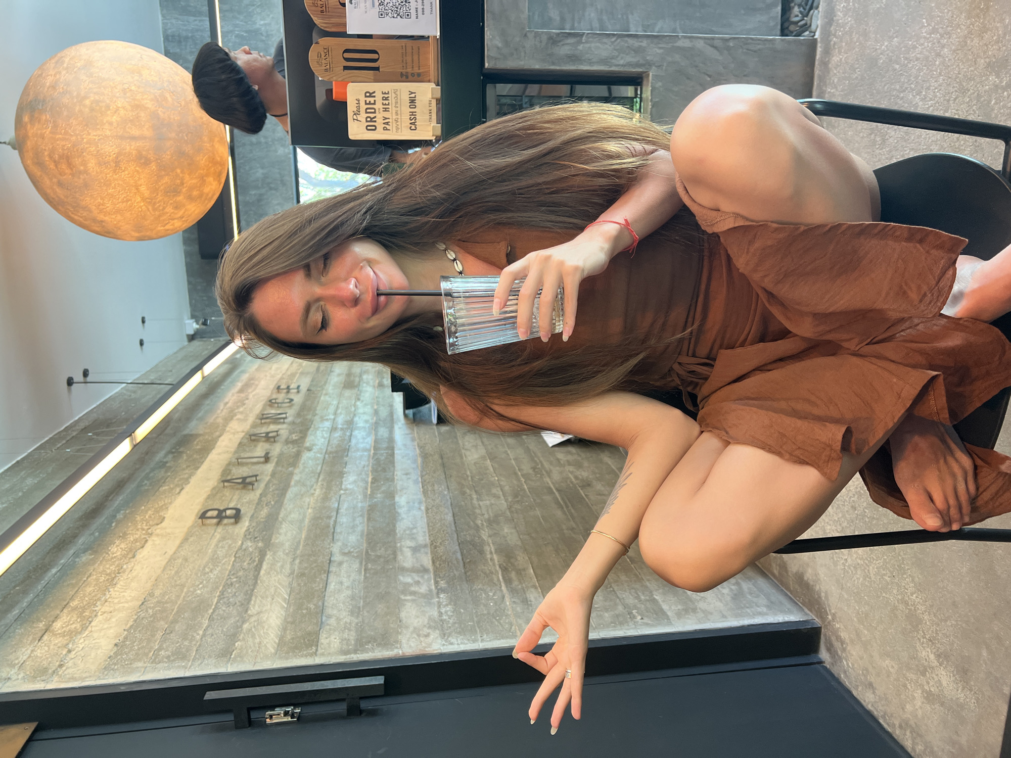 A young woman with long, brown hair is sitting on a chair in what appears to be a cafe or restaurant. She is wearing a brown dress and is holding a glass of water with a straw, from which she is drinking. Her eyes are closed and her mouth is slightly open, giving her a relaxed and perhaps content expression. A thin red string is tied around her left wrist, and a gold bracelet is visible on her right arm. She is also wearing a ring on her right ring finger.
The background includes a large, circular light fixture on the left, and a counter with various signs in the middle. One sign indicates "Please ORDER PAY HERE CASH ONLY". The word "BALANCE" is visible in large metal letters on a textured concrete wall behind her. The overall setting seems modern and industrial, with a concrete floor and wall. The lighting suggests it is daytime. The provided location context places this scene in Koh Phangan, Thailand.