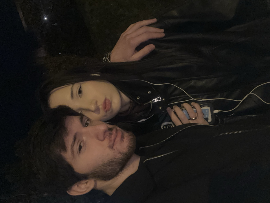 A young couple poses closely together at night. The man has dark hair and a beard. The woman, with long dark hair, is behind him. They both look at the camera. The woman wears a black leather jacket, and a hand with a watch is visible above her. A phone plugged into a charger is in her other hand. The man wears a black jacket. The lighting is dim, giving the scene a nighttime aesthetic.