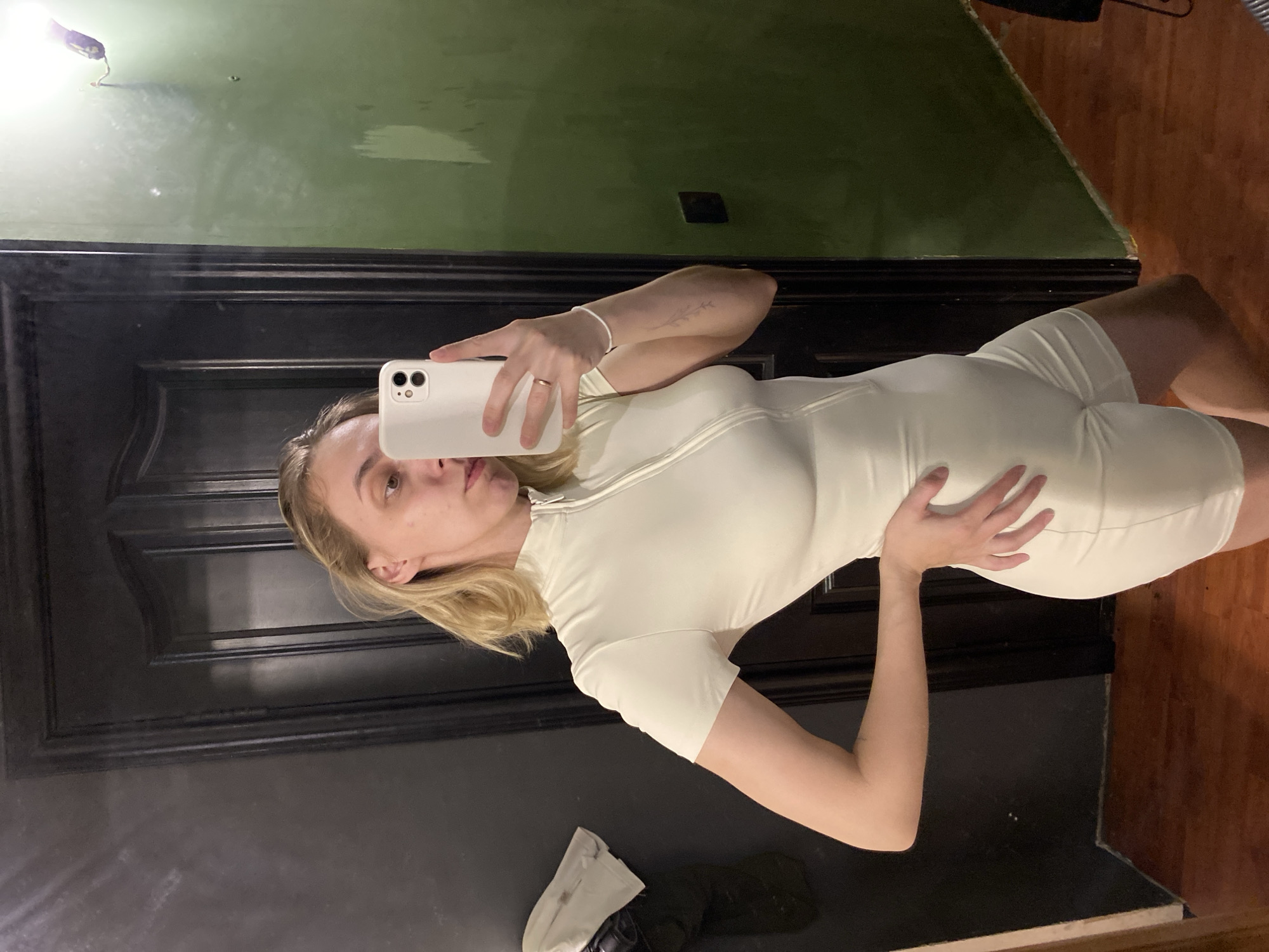 A woman with blonde hair is taking a selfie in front of a dark door. She is wearing a light-colored, form-fitting outfit. The background includes walls painted in shades of green and a wooden floor. The reflection suggests she may be in an indoor space. There is an object covered with a white cloth on the lower left. There is no confirmed location for Chaikovsky, Russia.