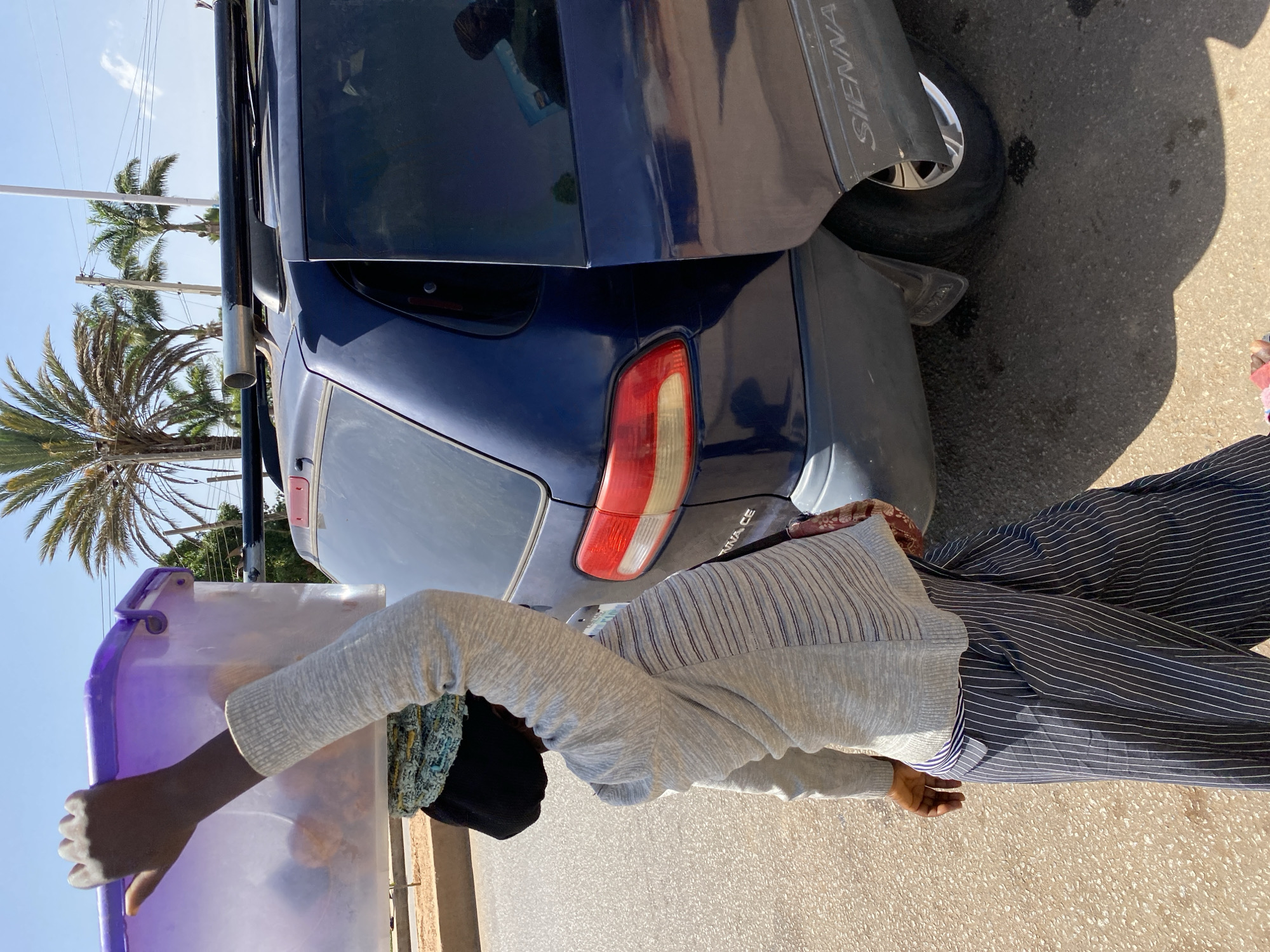 The scene shows a street in Bukuru, Nigeria, partially occupied by a dark blue Toyota Sienna minivan. A person wearing a grey sweater and striped pants is standing near the vehicle, holding a clear purple container. Palm trees and utility poles are visible in the background under a bright sky. The location is on paved asphalt.