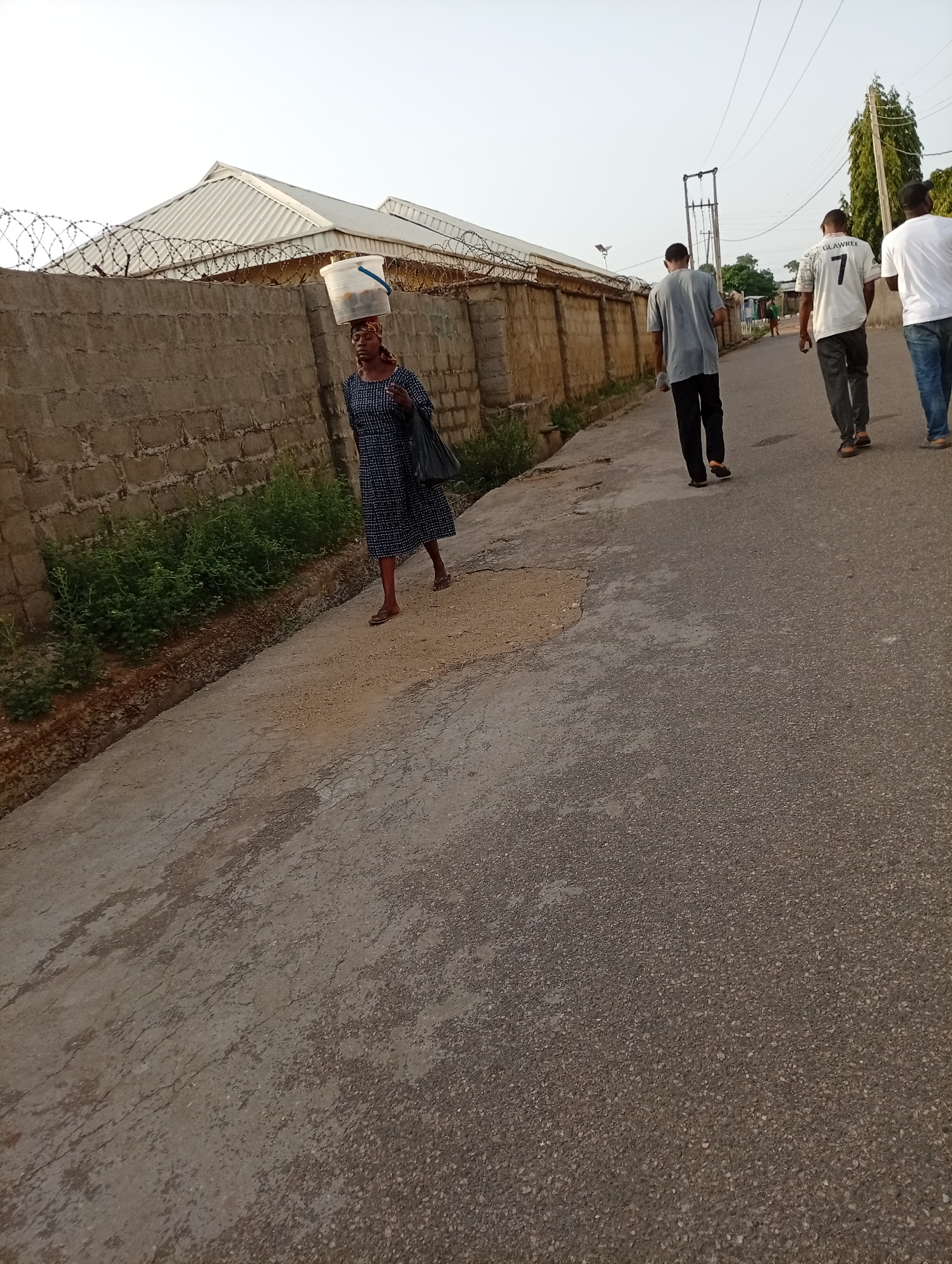 The photograph captures a street scene in Unknown, Unknown, showcasing an interplay of human activity and urban environment. A woman carrying a white bucket on her head is walking along the side of the road, a black bag hanging from her shoulder. She is wearing a dark, patterned dress and simple sandals.
Further down the street, three men are walking away from the camera. The man closest to the center is wearing a white jersey with the number 7 and the name "GLAWREE" on the back, while the others are wearing a gray and white t-shirt. They are all dressed in casual clothing.
The backdrop is defined by a tall, weathered wall topped with barbed wire, possibly enclosing a building with a metal roof. Greenery grows along the base of the wall. The street is paved and shows signs of wear and tear. Overhead power lines are visible, stretching across the frame. Tall trees stand in the distance. The lighting suggests it is daytime, with a slightly overcast sky.