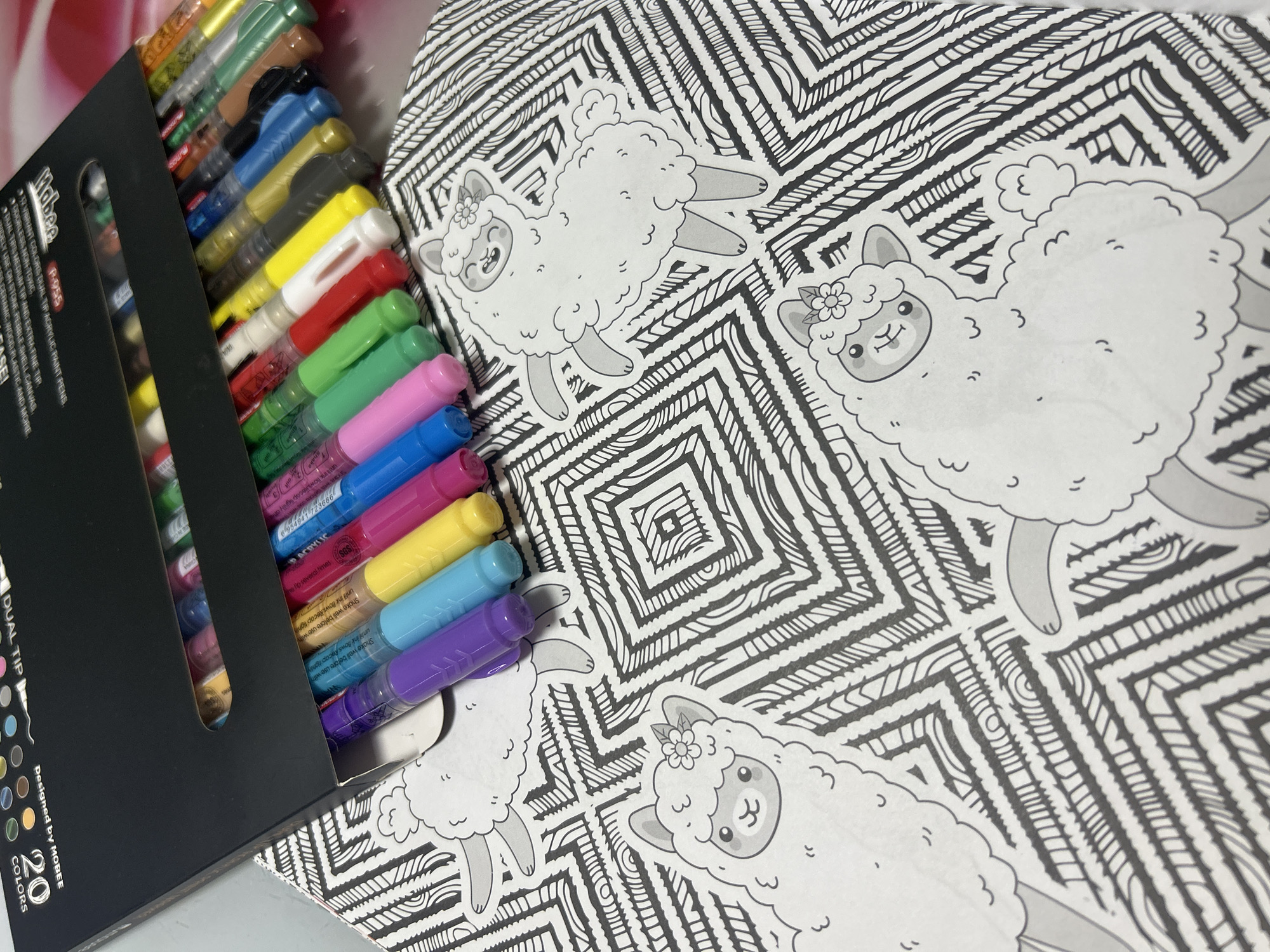 The image shows a box of 20 Mobee dual-tip acrylic paint pens, positioned on a coloring book page featuring line drawings of llamas within an intricate geometric pattern. The pens are arranged in the box with the caps visible, displaying a variety of colors including green, pink, blue, yellow, red, and metallic shades. The coloring book lies flat, showcasing the black and white illustrations. The environment appears to be indoors, with a neutral background. The overall focus is on the art supplies and the coloring book, suggesting a creative activity or hobby. There is no visible indication of Izhevsk, Russia.
