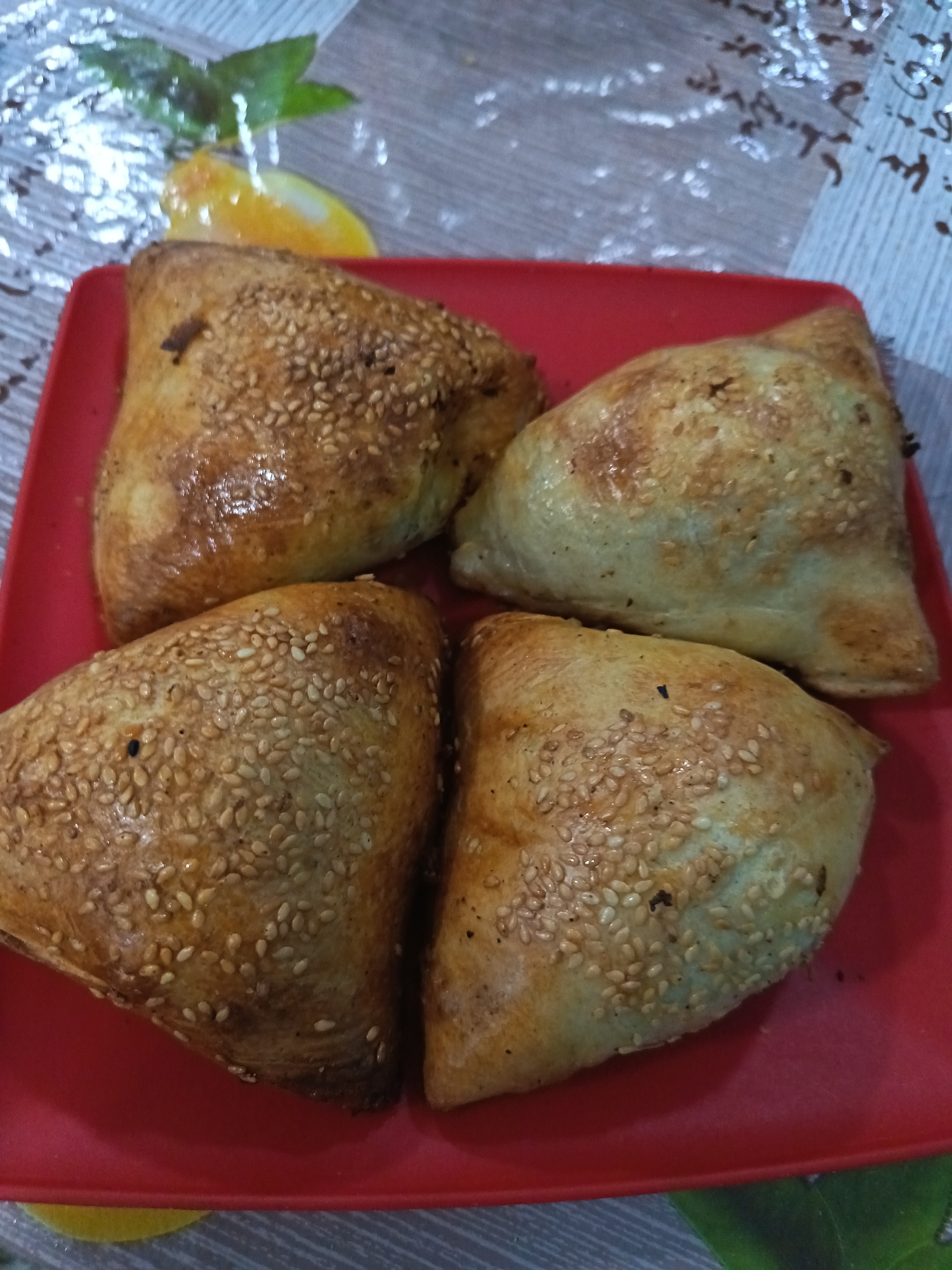 Four triangular baked pastries, possibly somsas, are arranged on a red rectangular plate. The pastries are golden brown and sprinkled with sesame seeds. The plate is set on a textured surface with a blurred background featuring a pattern that includes what appears to be a depiction of a lemon or orange slice and green leaves. The location is identified as Salar, Uzbekistan.