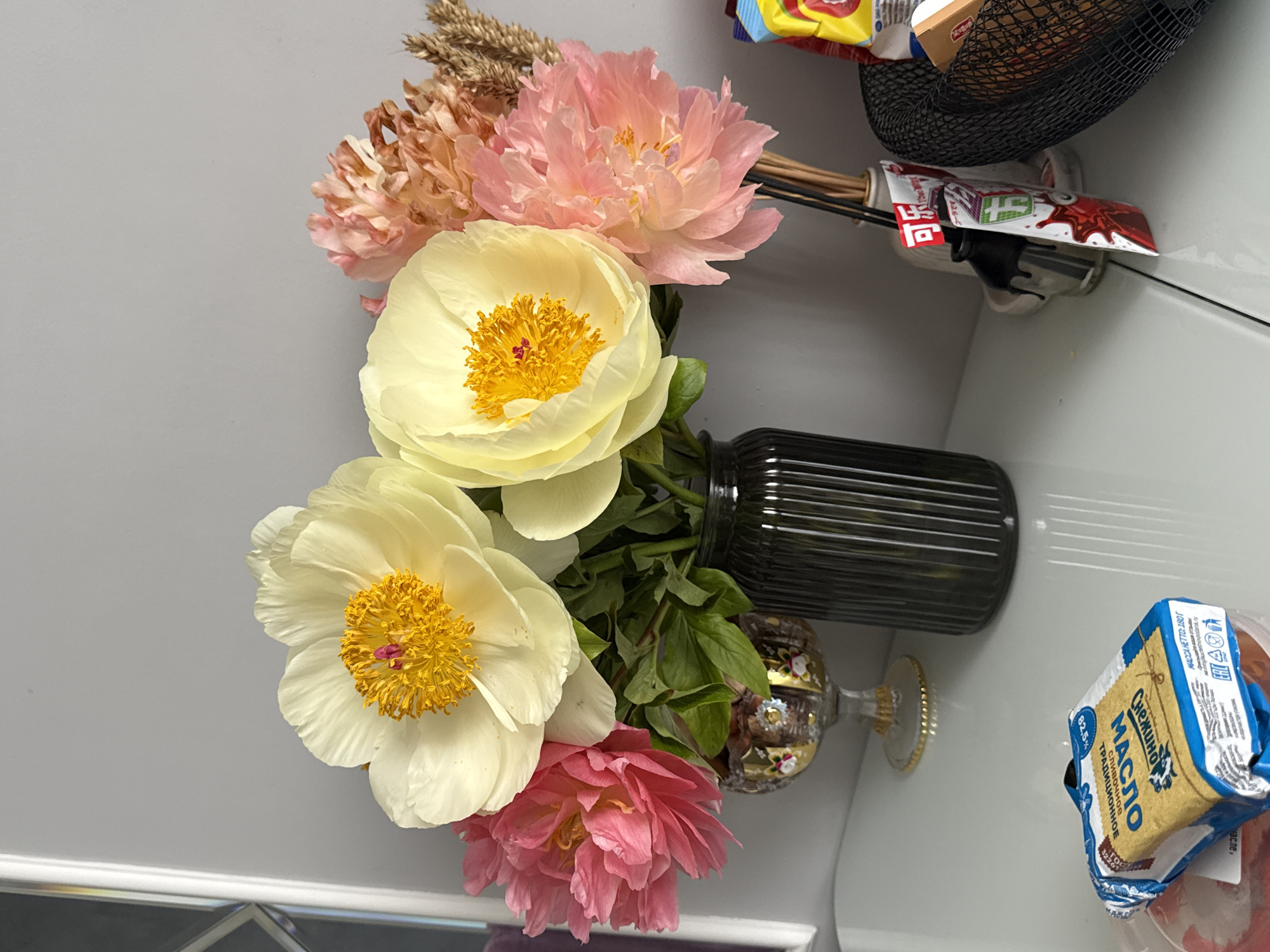 A still life depicts a bouquet of flowers arranged in a black ribbed vase, which appears to be placed on a white surface near other household items. The flowers consist of a mix of white and pink peonies, along with some dried wheat or ornamental grasses. The white peonies are prominently displayed, showcasing their bright yellow centers. Adjacent to the vase, on the right, is a partially visible blue and white package with Cyrillic script, possibly butter, and above the vase is a black mesh basket containing various items including a red and white tube of what appears to be food product. The background is a light gray, suggesting the scene is set against a wall or similar vertical surface.