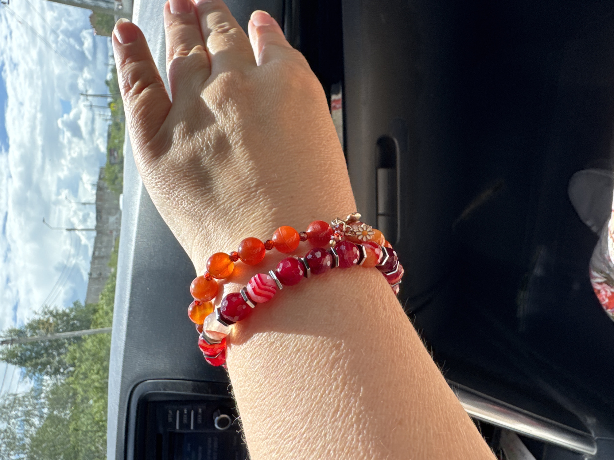 In Uchaly, Russia, a close-up shot captures a person's arm and hand resting on the interior of a car. Two bracelets adorn the wrist: one features faceted red and white agate beads with silver spacers, while the other is composed of smooth orange and red beads. The hand is partially visible, with fingers extended and a shadow cast from sunlight. The car's dark dashboard is in the foreground, and through the windshield, a slightly blurred view of trees and buildings against a cloudy sky is visible.