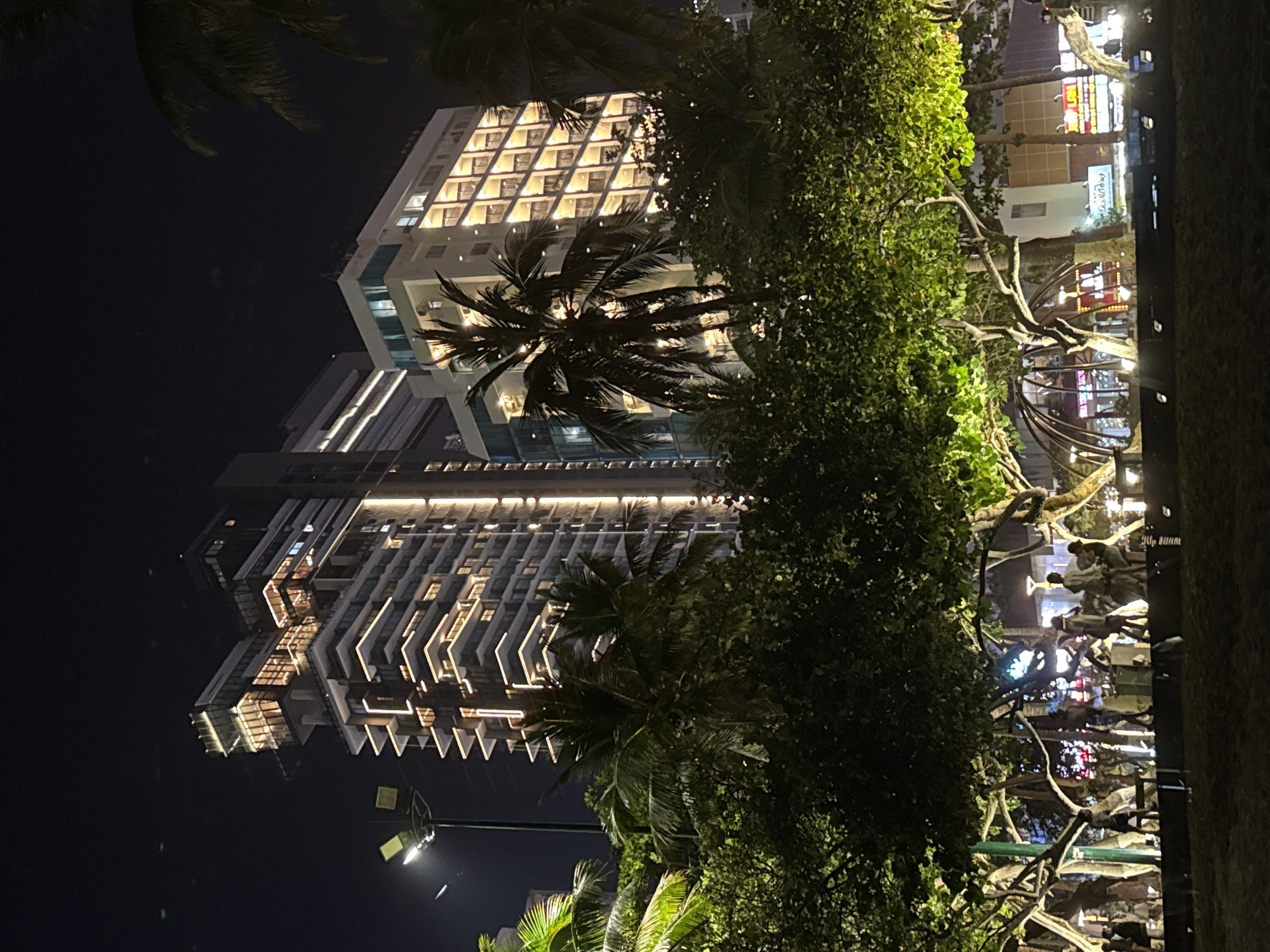 Captured at night, the image features two high-rise buildings in Unknown, Unknown. The building on the left exhibits a terraced design with illuminated balconies, while the building on the right displays a grid-like facade, also lit up. Palm trees are strategically placed in front of the buildings, adding a tropical aesthetic to the urban environment. On the right side of the image, there is a dense green area with trees that are adorned with decorative lighting. Pedestrians are seen strolling in the illuminated area next to the green space, suggesting a lively atmosphere. Streetlights provide additional illumination, enhancing the visibility of the scene. The photograph captures a blend of urban architecture and natural elements in the heart of the city.