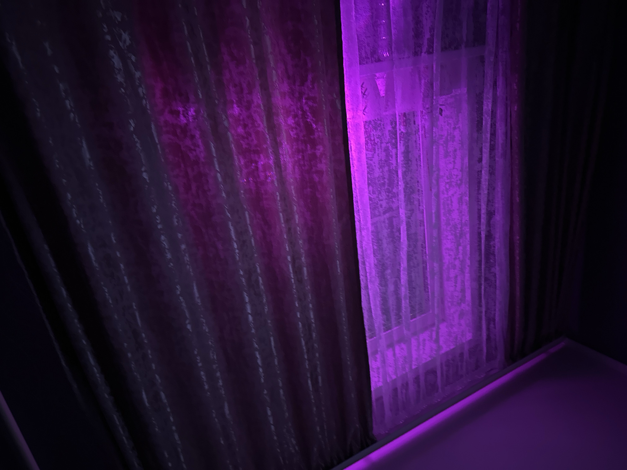 A dimly lit interior scene shows a window covered by sheer purple curtains and thicker, dark purple drapes. The lighting casts a vibrant purple glow on the sheer curtains, revealing the faint outline of a window frame behind them. The thicker drapes are textured and hang in vertical folds, also illuminated by the purple light from the left side of the frame, becoming progressively darker towards the right. The ceiling above is visible in the top left corner and is also lit with a purple hue. The scene appears to be indoors, possibly a bedroom or living space, with no people or specific objects clearly identifiable beyond the window treatments and ambient lighting. There is no information to associate this image with Podolsk, Russia.