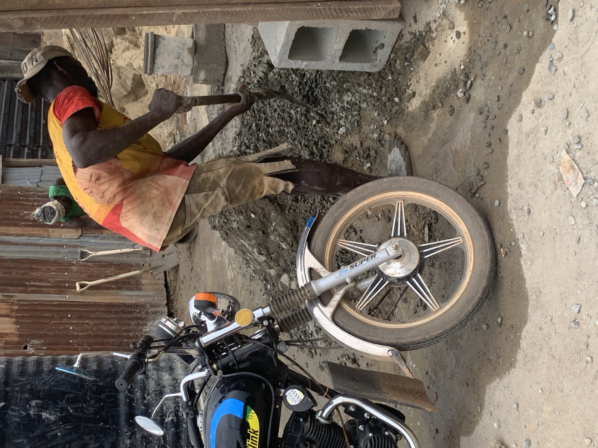 In Shagamu, Nigeria, a construction scene unfolds with a motorcycle prominently parked on the left. A man wearing a faded yellow and red shirt, tan shorts, and a cap is actively engaged in construction work, using a shovel to move a pile of gravel and cement. Behind him, another individual, partially visible, also appears to be involved in the construction process. The environment suggests an outdoor worksite with elements such as a corrugated metal wall, building materials like concrete blocks, and scattered debris on the ground. The presence of the motorcycle indicates potential transportation or a personal item at the site. The overall atmosphere is one of manual labor and ongoing construction in a possibly informal urban setting.