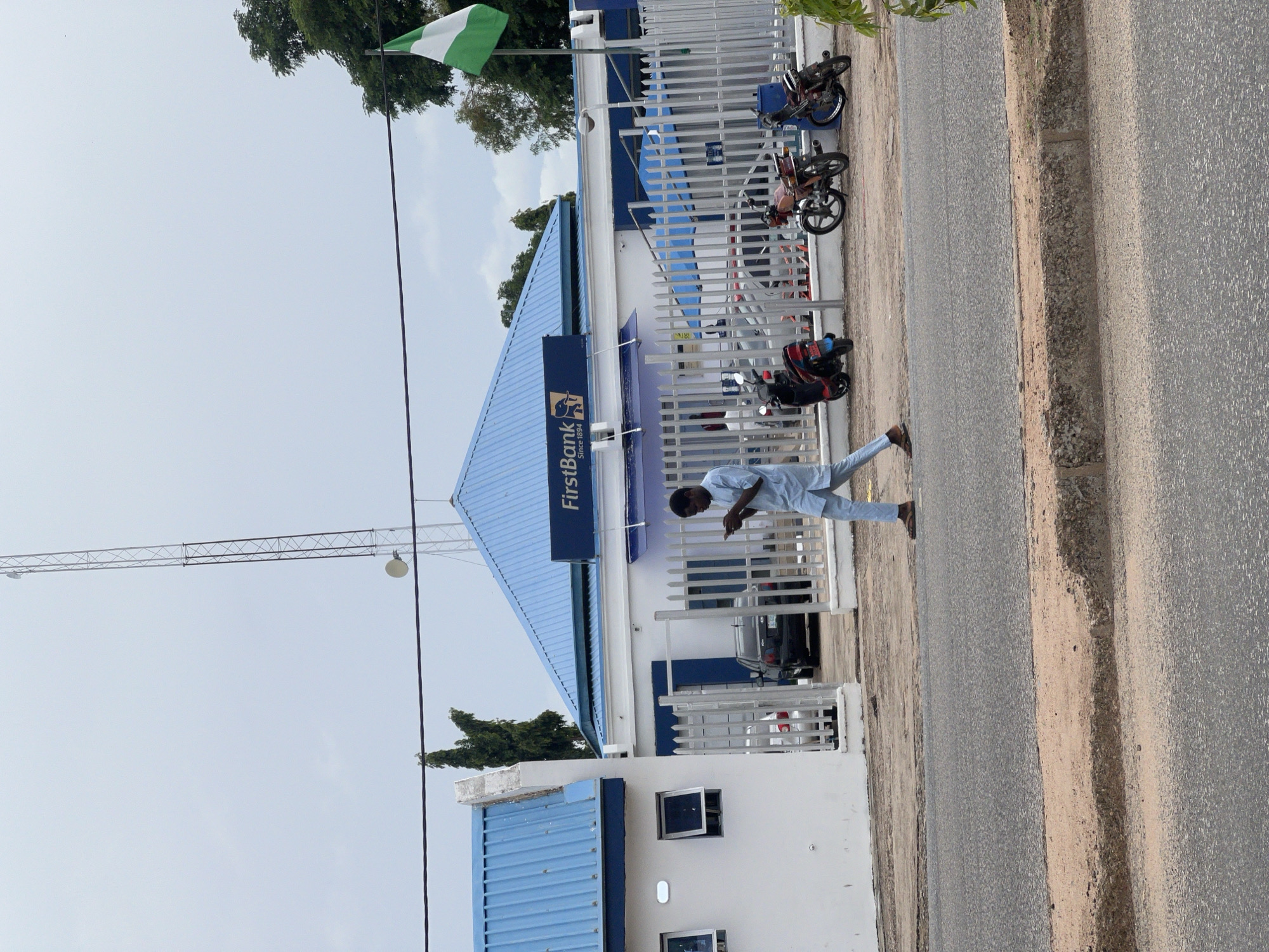 The image depicts the FirstBank building in Jalingo, Nigeria, under a cloudy sky. A man dressed in light blue traditional attire is walking across the foreground, looking at his mobile phone. To his right, a white metal fence encloses the bank's premises, where several motorcycles are parked, including a red one, a reddish-brown one, and a blue and black one. Beyond the motorcycles, a black car is visible, parked behind the fence. The bank building features a blue corrugated metal roof and white walls. A Nigerian flag, green and white, flies from a pole on the right side of the building. In the background, a tall telecommunications tower stands against the sky, and trees are visible to the left and right of the bank. A paved road with a sandy shoulder occupies the lower portion of the image.