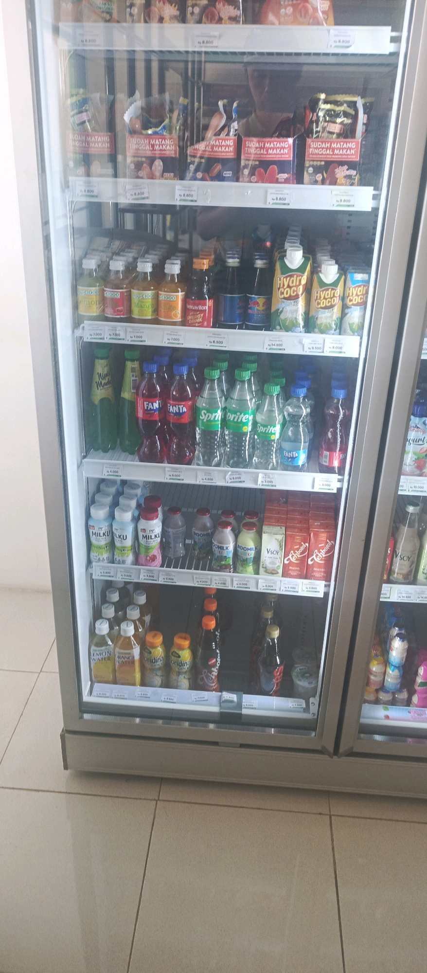 The image shows the interior of a commercial refrigerator in a convenience store, likely located in Lamongan, Indonesia. The refrigerator is filled with various beverages and food items, primarily drinks in bottles and cartons, and some packaged food items on the top shelves.
The top shelves display packaged food, possibly ice cream or frozen snacks, with labels that are partially visible and suggest prices in Indonesian Rupiah. Below this, multiple shelves are stocked with a variety of drinks. These include small bottles of flavored drinks, energy drinks like Red Bull, and cartons of coconut water. Further down, there are rows of larger plastic bottles containing carbonated drinks such as Sprite and Fanta, and other flavored beverages. The bottom shelves contain milk-based drinks in bottles, some cartons labeled "Teh Botol," and what appear to be yogurt cups or small desserts.
The refrigerator doors are transparent, allowing a view of the products inside. A reflection of a person, likely an employee or customer, can be seen in the glass on the upper shelves, obscuring some of the products. The overall lighting is cool, typical of refrigerator interiors. The floor tiles are visible in the foreground, suggesting a clean and tiled environment typical of a retail space. The specific location is identified as Lamongan, Indonesia, implying the context of a local Indonesian market or store.