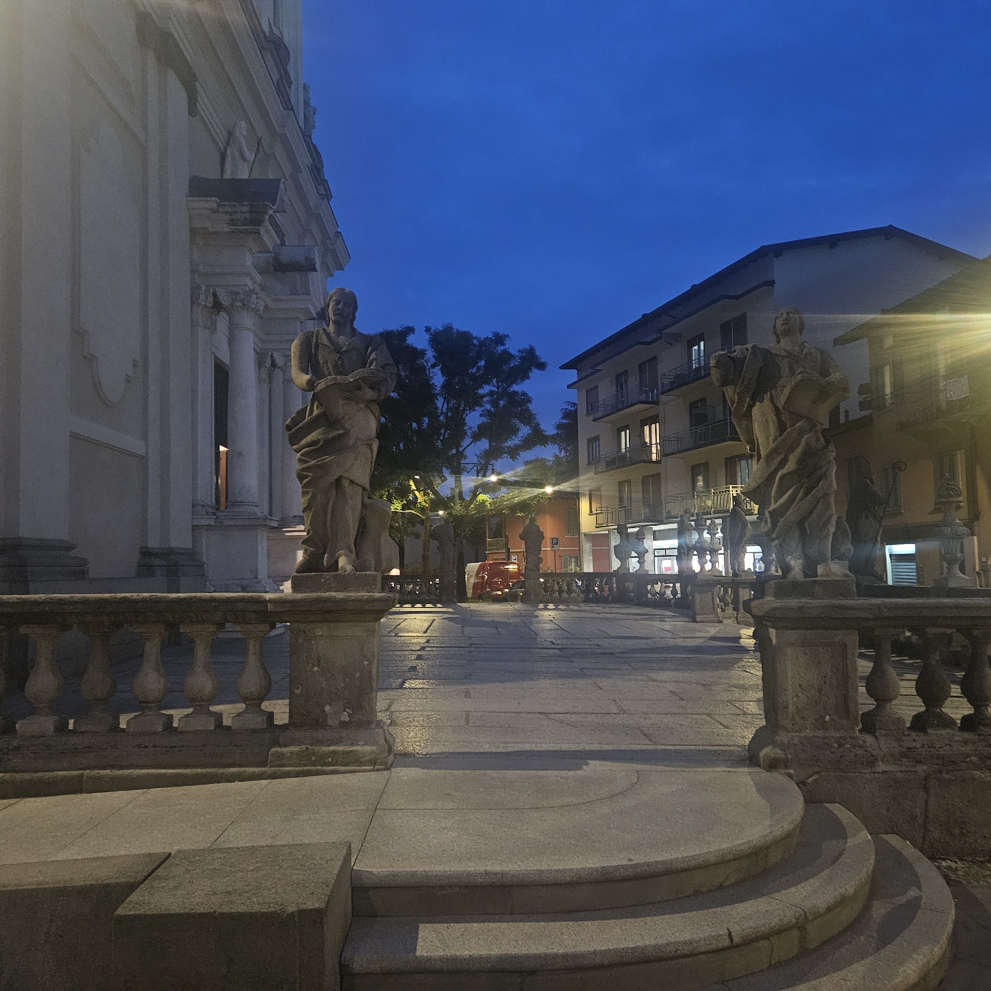 This is a view of the Piazza della Liberazione in Stezzano, Italy, at night. In the foreground, stone steps lead up to a wide, paved plaza with a stone balustrade on either side. Statues stand atop the balustrade on both sides. To the left is the facade of a grand building with columns. In the distance, buildings line the plaza, illuminated by streetlights and interior lights from shops. A red vehicle is visible in the background. The sky is a deep blue, suggesting twilight or nightfall.