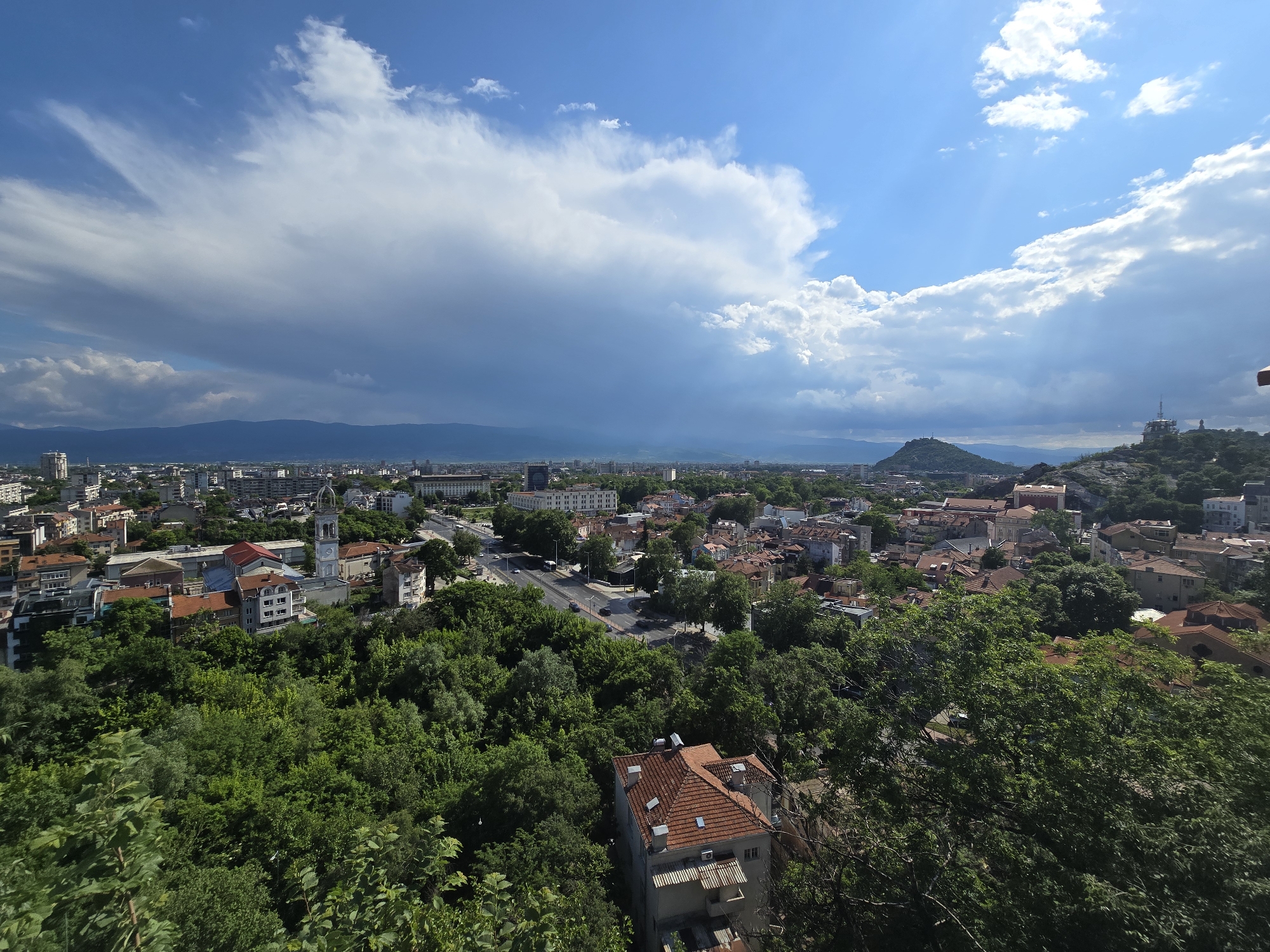 The image depicts a cityscape under a partly cloudy sky. The foreground is filled with dense green trees, partially obscuring buildings with red-tiled roofs. A road runs through the city. A clock tower is visible amongst the buildings. In the background, a mountain range is visible on the horizon. The city is Unknown, Unknown.