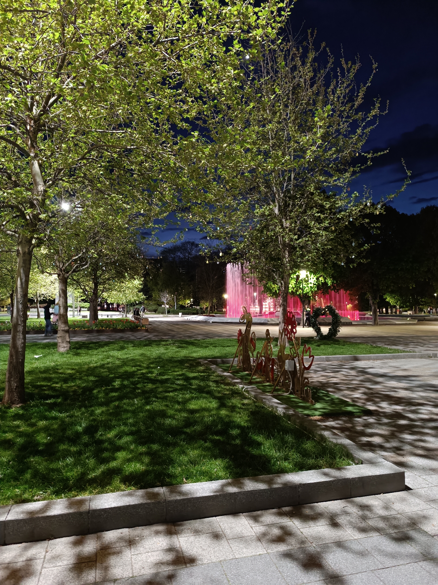 The image shows a park at night. The location is Unknown, Unknown. In the foreground, there is a lawn of green grass bordered by a gray concrete edge. Several thin trees are scattered around the lawn, their leaves illuminated by lights, possibly streetlights.
To the right of the lawn, there is a paved walkway with shadow patterns from the trees. Next to the lawn, there are decorative metal art figures that look like animals.
In the background, there are pink illuminated fountains. People are sparsely scattered in the park. Beyond the fountains, more trees and lights can be seen in the distance. The sky is dark, suggesting it is nighttime.