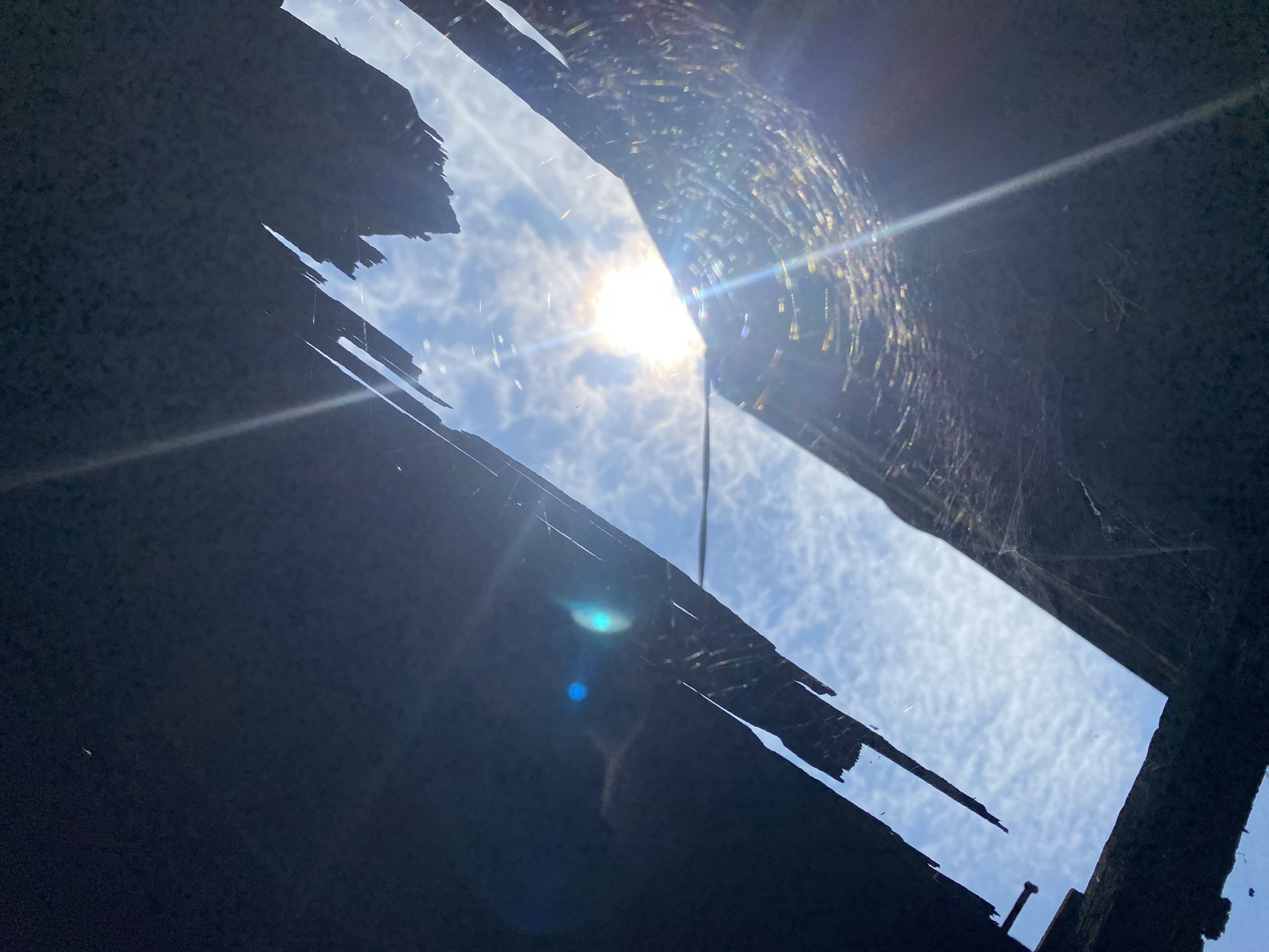 The image captures a view looking upwards through a damaged roof, revealing a bright sky with scattered clouds. The sun is a prominent feature, casting strong lens flares and illuminating a delicate spiderweb strung between the broken roof materials. The roof itself appears to be made of dark, possibly charred or weathered material, with irregular, jagged edges. The scene suggests an abandoned or damaged structure, possibly in a less developed area, though no specific landmarks or people are visible to definitively place it within Banjarmasin, Indonesia. The overall impression is one of decay and natural elements reclaiming a man-made space.