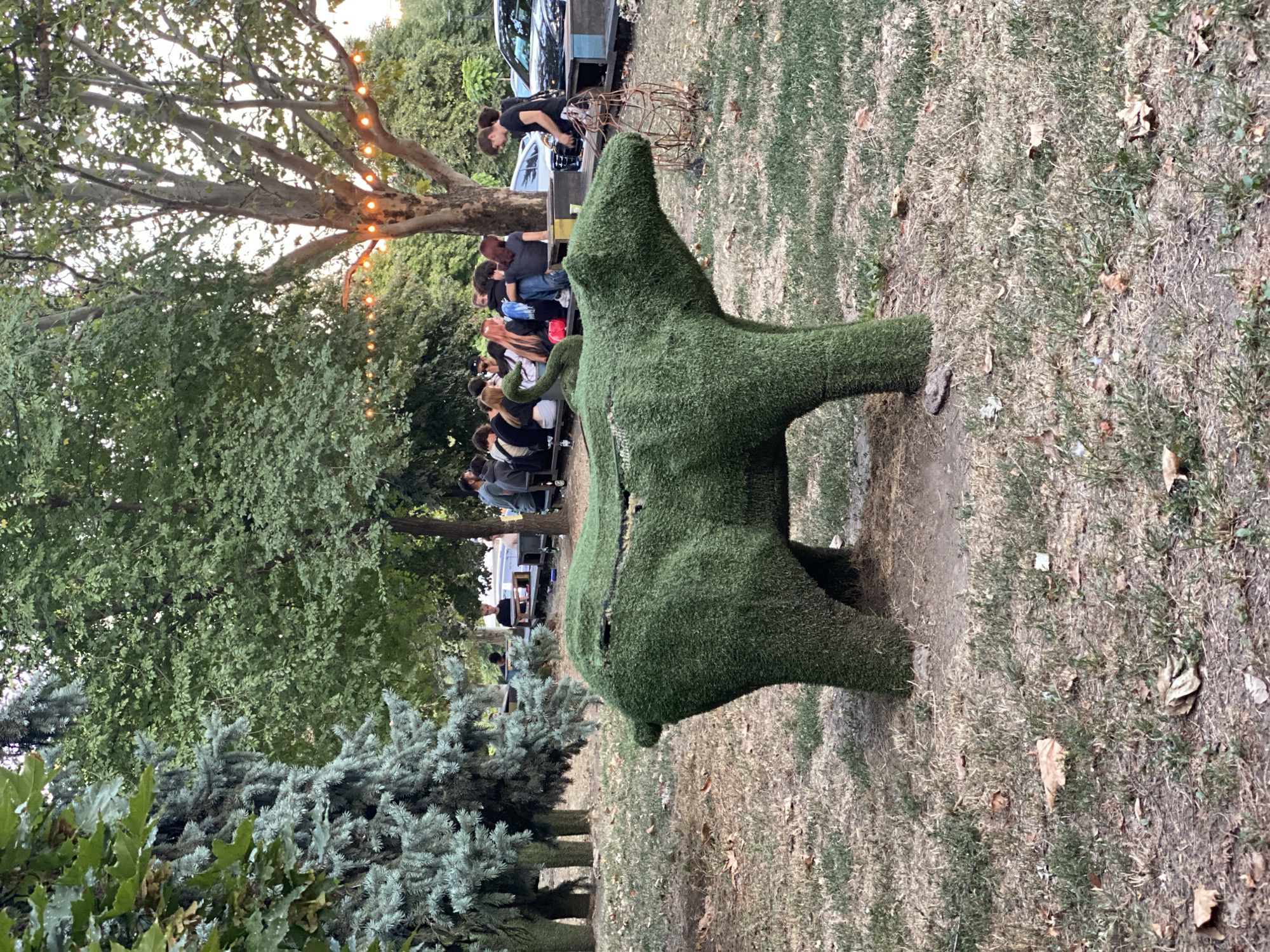 The image depicts an outdoor gathering in Chișinău, Moldova, likely in a park or beer garden setting. In the foreground, a large, grass-covered sculpture shaped like a bear is positioned on dry, patchy grass. Behind the bear sculpture, a group of people are seated at tables and on benches under a tree adorned with string lights. The background shows more trees and what appears to be a road with vehicles, suggesting an urban park or recreational area. The overall atmosphere appears relaxed and social, with individuals engaged in conversation and enjoying the outdoor ambiance. The presence of lush greenery, string lights, and casual seating arrangements points towards a pleasant evening or late afternoon scene.