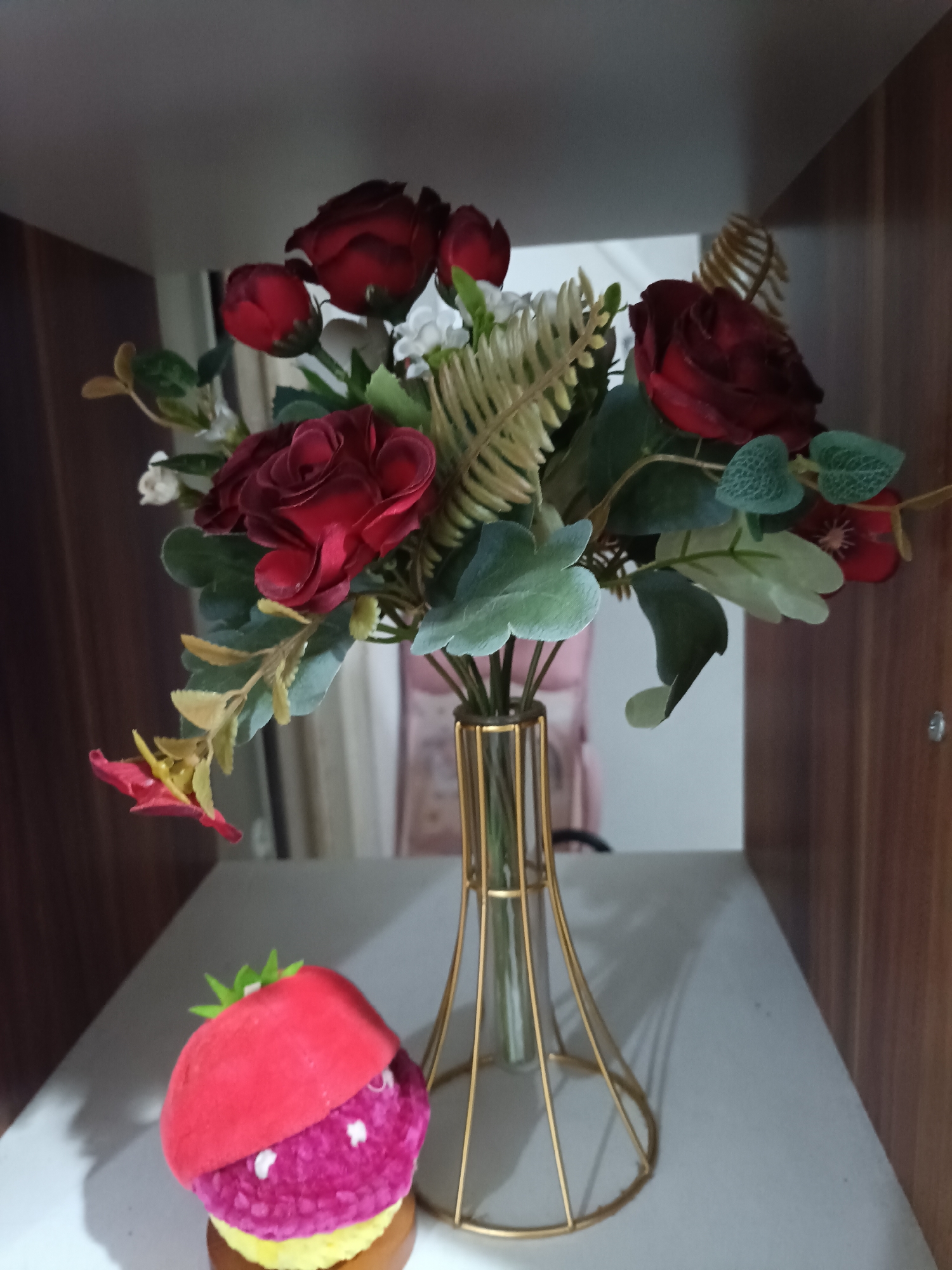 This media file features a still life arrangement on a shelf. The primary subjects are a bouquet of artificial red roses and greenery in a modern, gold wire vase. A small, decorative strawberry-shaped object sits in front of the vase. The scene is set within a shelf unit, with wooden paneling visible on the right side. The lighting suggests an indoor environment, possibly a home or office. There is no discernible activity or event taking place, as it is a static arrangement. Notable details include the textured appearance of the faux flowers and the strawberry decoration, which has a bright red top and a purple, fuzzy body with white dots. The setting appears to be a well-lit interior, likely during the daytime given the brightness. There is no visible text in the image. The location context of West Jakarta, Indonesia, is noted but not visually apparent in the image itself.