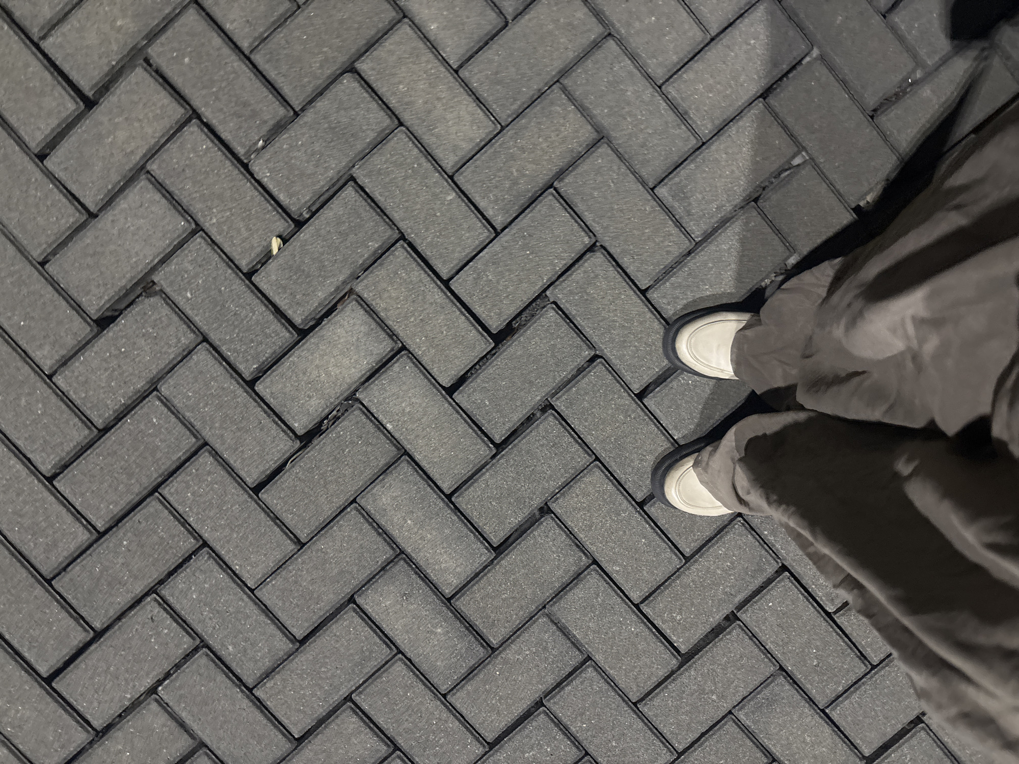 This media file shows a top-down view of a person's feet and legs standing on a dark gray paver brick pathway arranged in a herringbone pattern. The person is wearing light-colored shoes with dark soles and long, loose-fitting dark pants that obscure most of their body. The pathway is outdoors, and the lighting suggests it may be evening or nighttime, with shadows cast by the person. The bricks appear somewhat rough in texture. The scene is set in Babakan Madang, Indonesia. No specific activity is depicted beyond standing, and no text is visible.