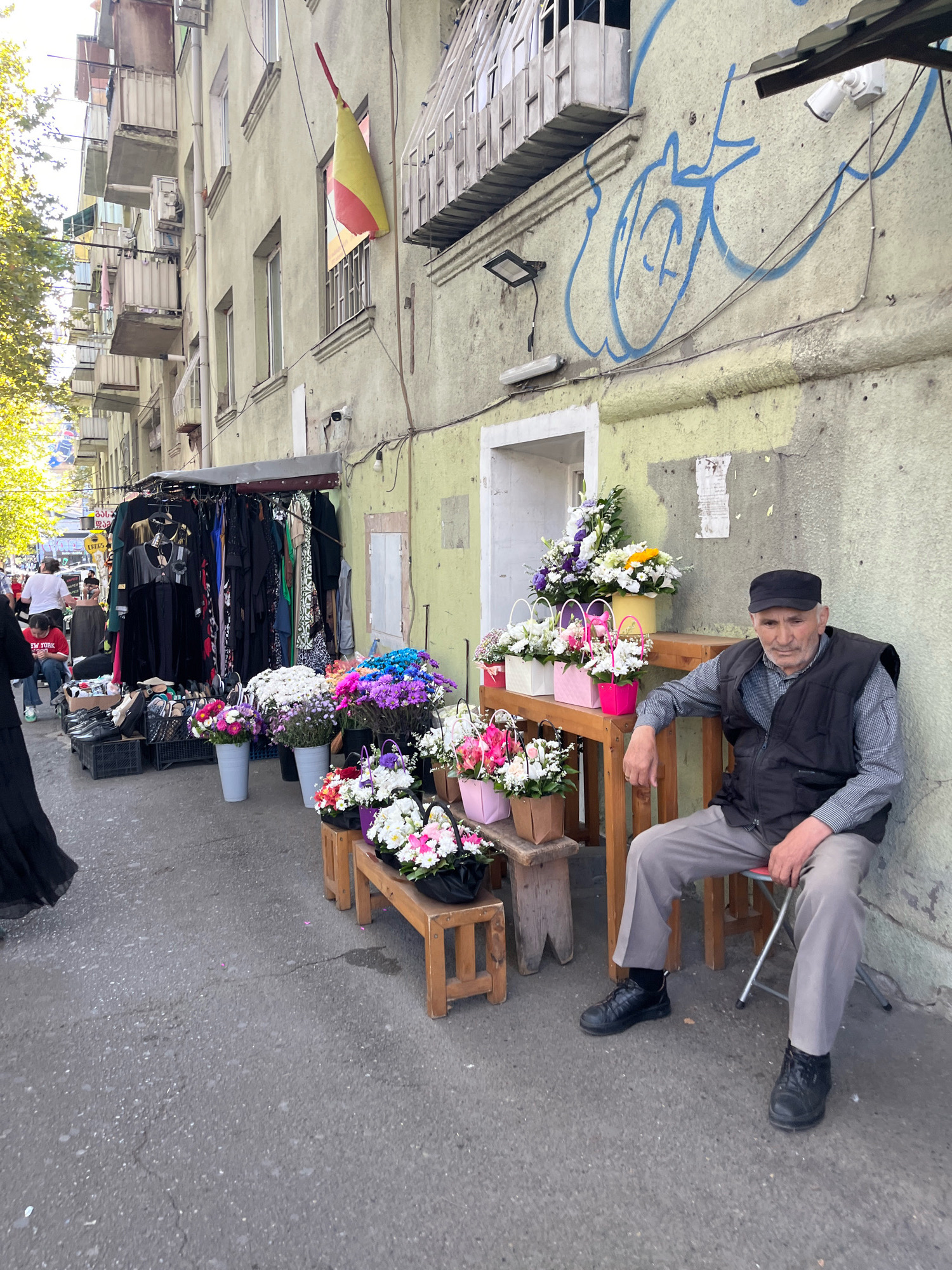 The image depicts a street scene in Tbilisi, Georgia, featuring an outdoor market stall with clothing and flowers. An elderly man, wearing a black vest over a checkered shirt and a black cap, sits on a foldable stool next to the flower stall. He is looking towards the camera. In the background, a building with balconies and graffiti is visible. The market stall itself displays a variety of clothing items on hangers and an assortment of colorful flowers in buckets and baskets. Several people are seen walking in the background, engaged in shopping or passing by. The ground is paved with asphalt, showing some wear and tear. The overall impression is that of a bustling urban environment during daylight hours.