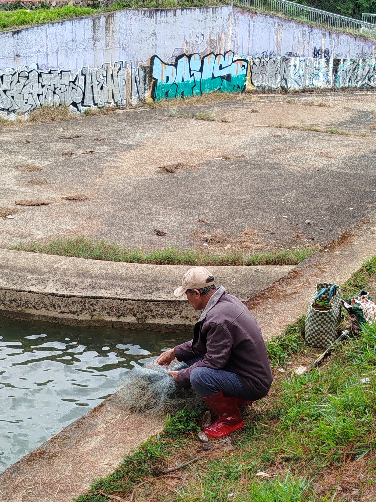 In Da Lat, Vietnam, an individual wearing a tan baseball cap, a dark jacket, blue jeans, and red rubber boots kneels by a concrete water feature. They are holding a cast net. Beside them on the grassy bank are two woven bags and a fishing rod. The water feature is lined with concrete, and the bank is covered in green grass and scattered leaves. In the background, a large concrete wall is covered in graffiti. The ground is a mix of concrete and dirt.