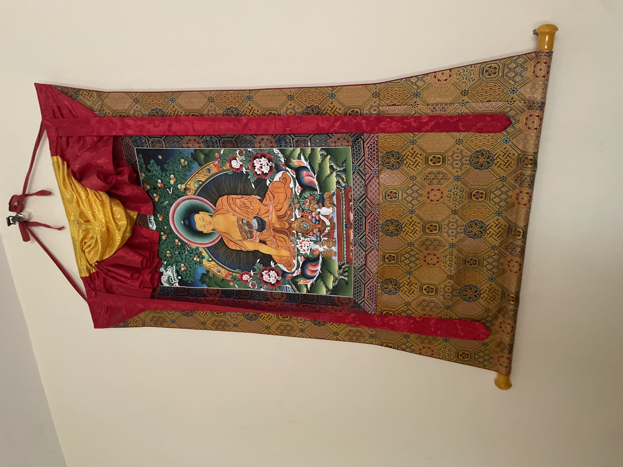 A Thangka, a traditional Tibetan Buddhist painting, is prominently displayed, hanging vertically against a plain, light-colored wall. The central artwork within the Thangka depicts a seated Buddha figure, rendered in warm orange and golden tones. The Buddha is shown in a meditative posture, with one hand resting in the lap and the other gently touching the earth, a gesture commonly associated with the Bhumisparsha Mudra. A radiant halo and an aureole encircle the Buddha's head and body. The background of the painting features lush green foliage, possibly a landscape with trees bearing fruits or flowers, and hints of a blue sky.
The painting is mounted on a larger textile scroll made of a rich, golden-brown brocade fabric. This fabric is intricately patterned with geometric and stylized floral motifs in shades of blue, red, and gold, creating a luxurious frame for the central image. Vibrant red silk borders frame the painting on all sides, extending upwards and downwards. At the top of the Thangka, draped over the red borders, are folds of bright red and golden-yellow silk, forming decorative elements. The entire Thangka is suspended from a horizontal wooden dowel at the top, and a similar dowel, with yellow finials, is partially visible at the bottom, providing weight and structure. No activity is taking place, and no visible text is present in the image.