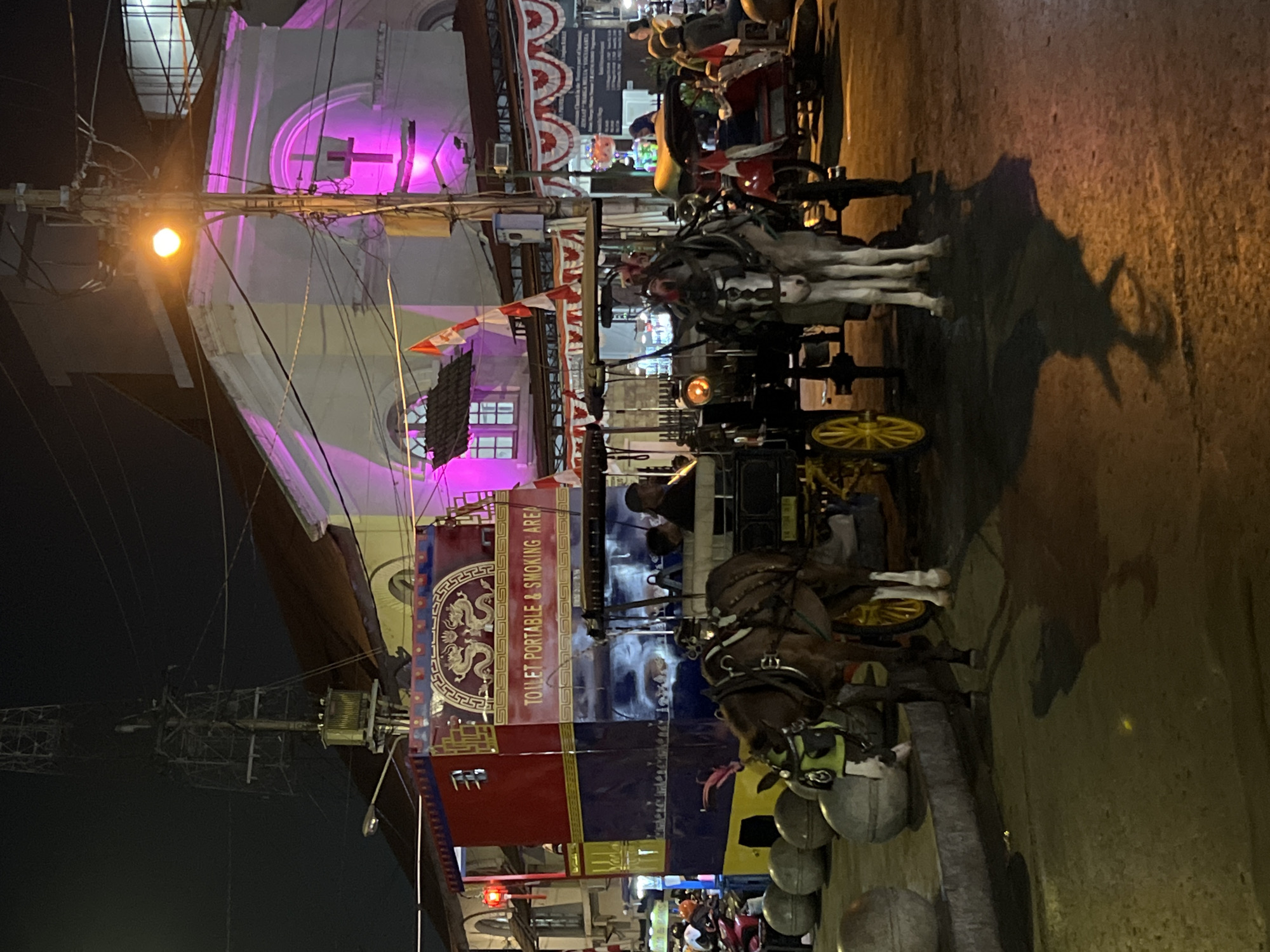 The image captures a nighttime scene in Malioboro Street, Yogyakarta, Indonesia, characterized by illuminated buildings and street life. In the foreground, two horse-drawn carriages, known as "andong," are parked. The carriage on the left is occupied by two individuals, with a dark-haired person in the driver's seat. A brown horse pulls this carriage, adorned with colorful harness and decorative elements. The carriage on the right is also occupied by a few people and is pulled by a white and grey horse. Behind the carriages, a large, multi-colored portable toilet and smoking area structure is visible, decorated with dragon motifs and text that reads "TOILET PORTABLE & SMOKING AREA." The building behind this structure is illuminated with vibrant pink lights, highlighting its architectural features, including a large cross, suggesting it might be a church or a religious building. The street itself is paved, and several large, rounded concrete bollards are present on the left side of the frame, demarcating an area. Various other elements like streetlights, overhead wires, and distant motorcycles contribute to the urban atmosphere. The overall impression is of a lively commercial and tourist area in Yogyakarta at night.