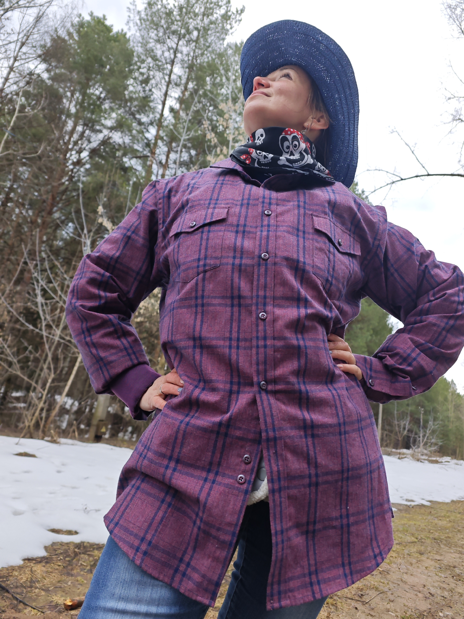 A woman stands outdoors in a landscape with melting snow and trees, likely in a wooded area. She is wearing jeans, a purple and blue plaid button-down shirt, a black bandana with a skull print around her neck, and a dark blue woven hat. Her hands are on her hips, and she is looking upward. The ground is a mix of dirt, grass, and patches of snow. Bare trees are visible in the background. The location is in the city of Chaikovsky, Russia.