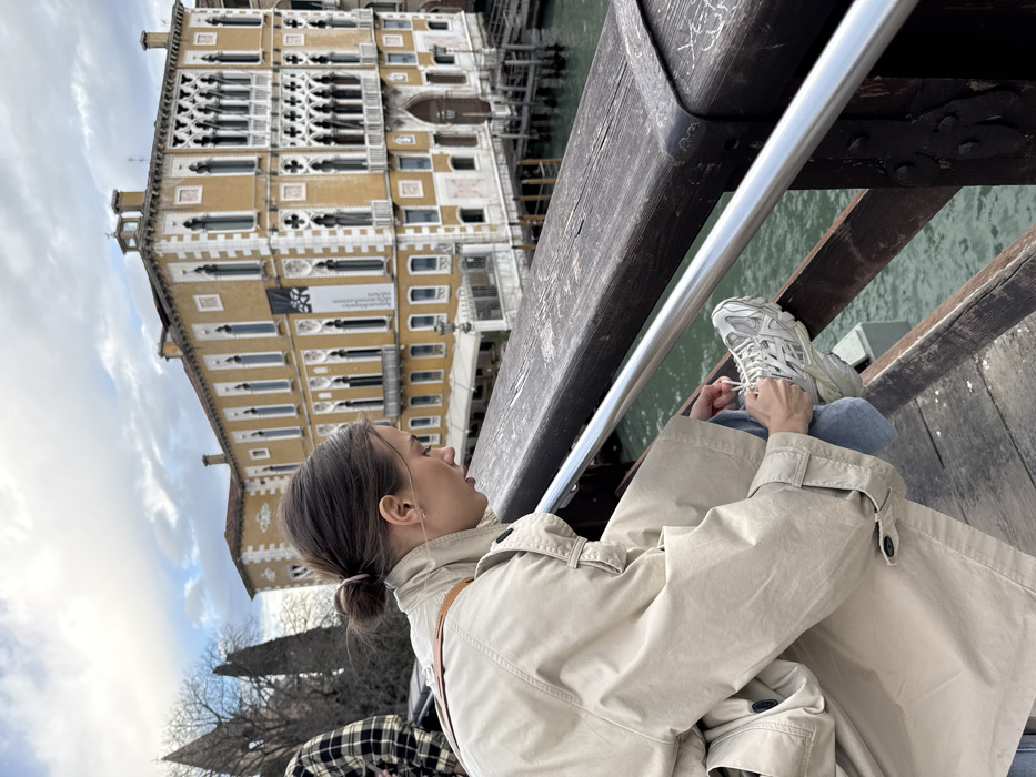 A female individual is depicted in the foreground on a wooden pedestrian bridge or platform overlooking the Grand Canal in Venice, Italy. She is bent over, tying the laces of her right light gray athletic shoe, which is propped against the dark brown wooden railing. She wears a light beige trench coat, light blue jeans, and her dark hair is gathered in a bun. A light brown strap, possibly from a bag, is visible over her right shoulder. The wooden railing, topped with a metallic silver handrail, shows signs of weathering and minor superficial markings.
In the midground, across the Grand Canal, stands the Palazzo Giustinian, a prominent multi-story building characterized by its ochre facade with white decorative trim and numerous ornate Gothic-style arched windows. A black vertical banner with a white logo and text, identifying it as the European School of Economics (ESE), hangs on the building's facade. The greenish-blue water of the Grand Canal is visible beneath the bridge, with wooden structures and docks extending from the palazzo's base. The background sky is overcast with patches of brighter light. On the far left, a portion of another person wearing a black and beige plaid shirt is partially visible, behind some bare trees.