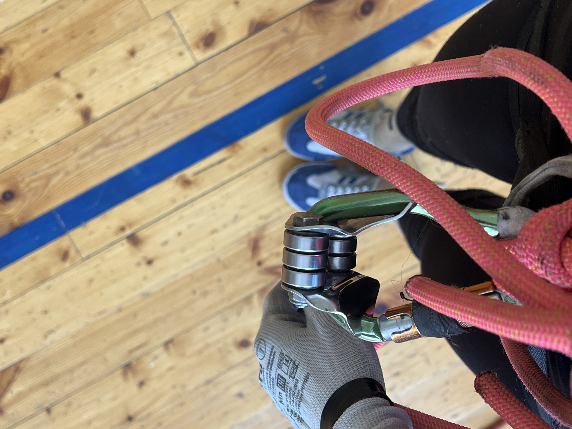 A person wearing gray gloves is holding a silver climbing ascender attached to a green carabiner and a pink rope. The scene appears to be indoors on a light wood floor with a blue stripe painted on it. In the background are a pair of blue and gray sneakers. No specific details link this image to the city of Chaikovsky, Russia.