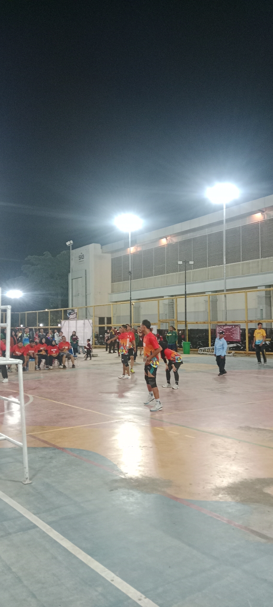 The image shows a volleyball game at night on an outdoor court in Tuban, Indonesia. The court has a red and yellow surface with white boundary lines. There are several people on the court, including players in red and orange jerseys and spectators sitting on benches. The court is illuminated by bright floodlights mounted on tall poles. In the background, there is a building with a white facade and a grated wall. The sky is dark, indicating it is nighttime.