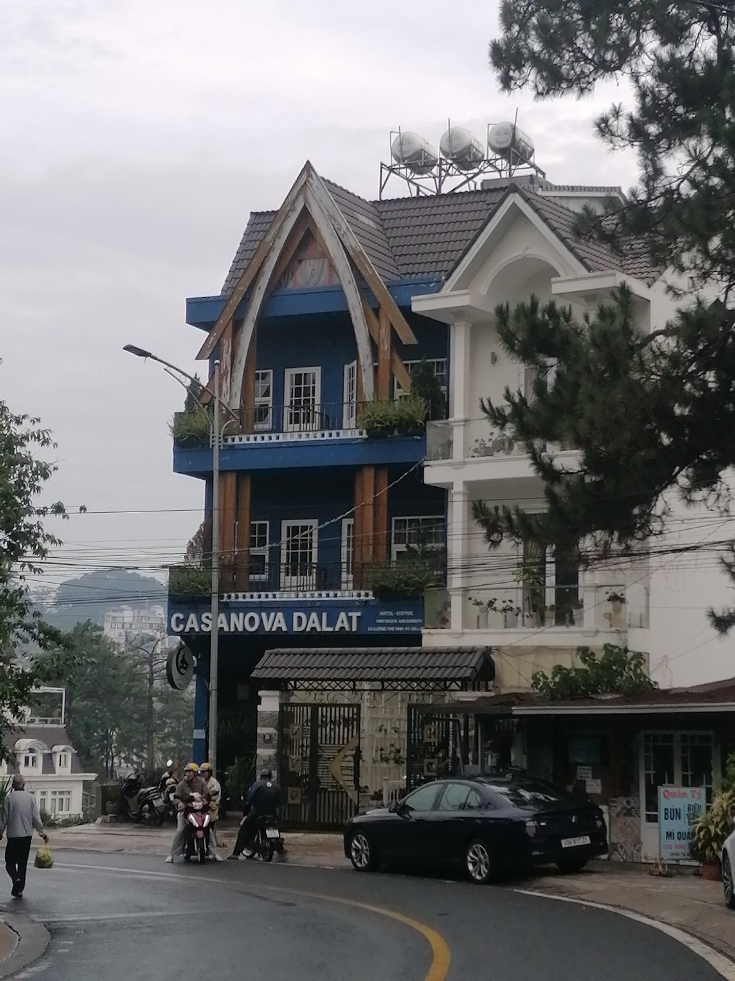 The image shows the exterior of Casanova Dalat, a hotel in Da Lat, Vietnam. The building is three stories tall, with blue and white sections, distinctive arched wooden details on the blue part, and tanks on the roof. In front of the building, there's a street scene with a black car parked, and several people on motorbikes. A man is walking on the street in the foreground. There is also a small restaurant next to the Hotel. The weather seems overcast. In the background, there is a view of the hilly landscape and buildings of Da Lat.