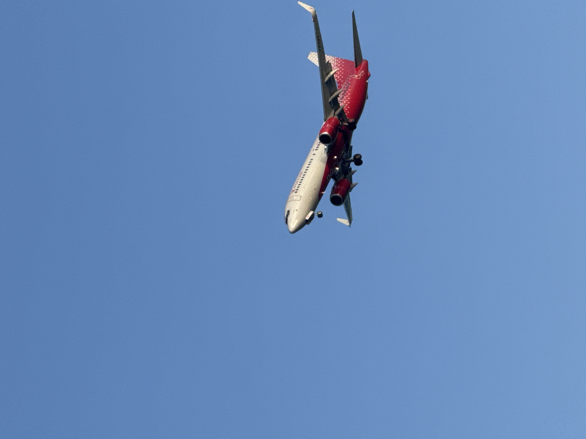 A low-angle shot of a commercial airliner, painted red and white, flying upside down against a clear blue sky. The plane's landing gear is deployed. The location is not discernible from the image. The subject is the airplane. There are no visible people.