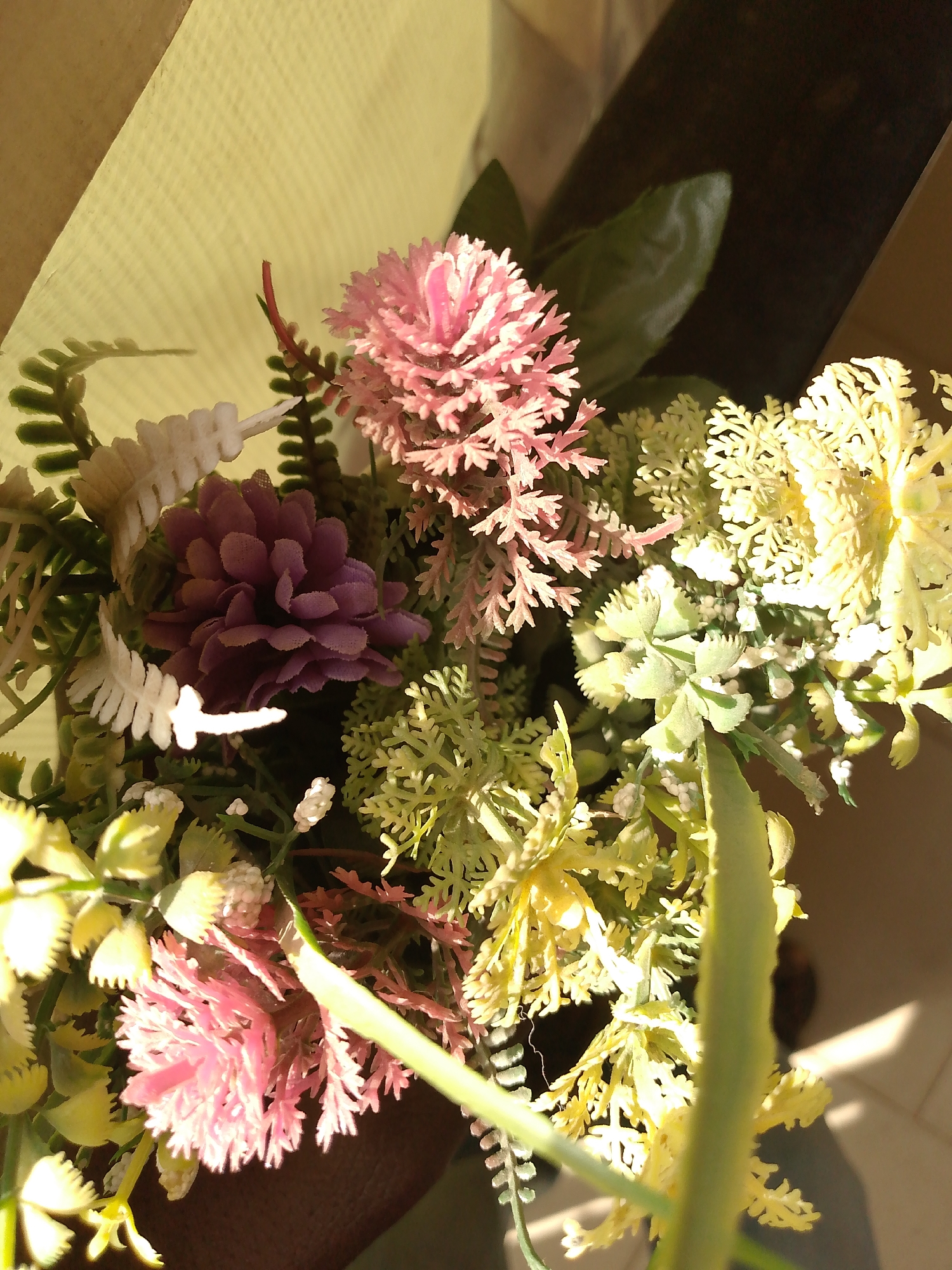 A bouquet of artificial flowers in various colors is presented in a brown paper bag. The flowers include pink, purple, white, and yellow varieties. The arrangement is brightly lit, possibly by sunlight. The location in Bukuru, Nigeria, is not verifiable from the image. No people are visible.