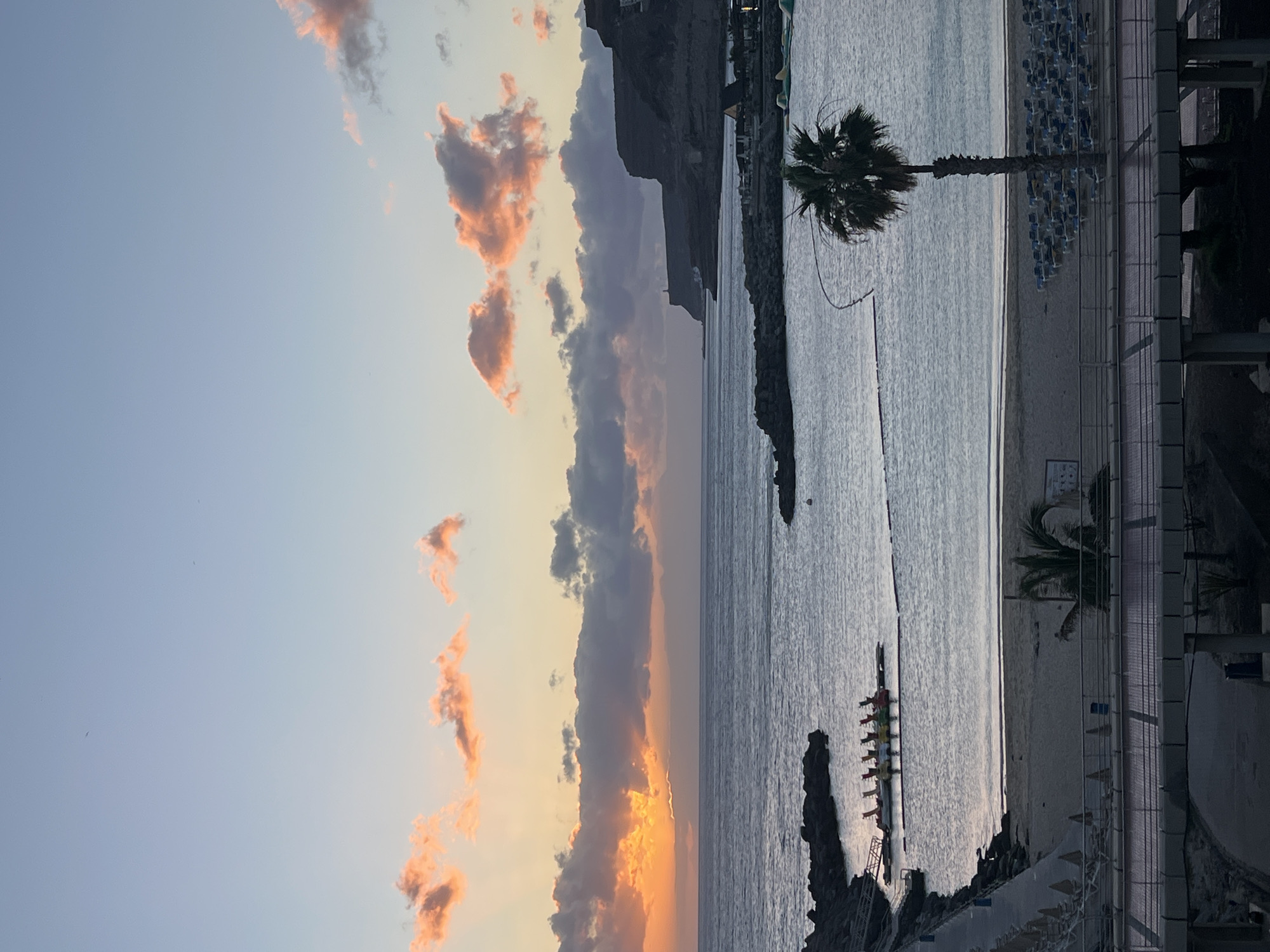 The image depicts a coastal scene in Mogán, Spain, at what appears to be sunset or sunrise. The sky shows hues of blue and orange, with scattered clouds tinged with pink. A strip of land extends from the left side, consisting of dark rocks and vegetation. The sea occupies the central part of the frame, reflecting the light and creating a shimmering surface. A palm tree stands prominently on a sandy beach. In the lower right, a walkway or structure is visible. On the left side, a boat with several occupants is near the shoreline, along with many beach chairs on the beach.