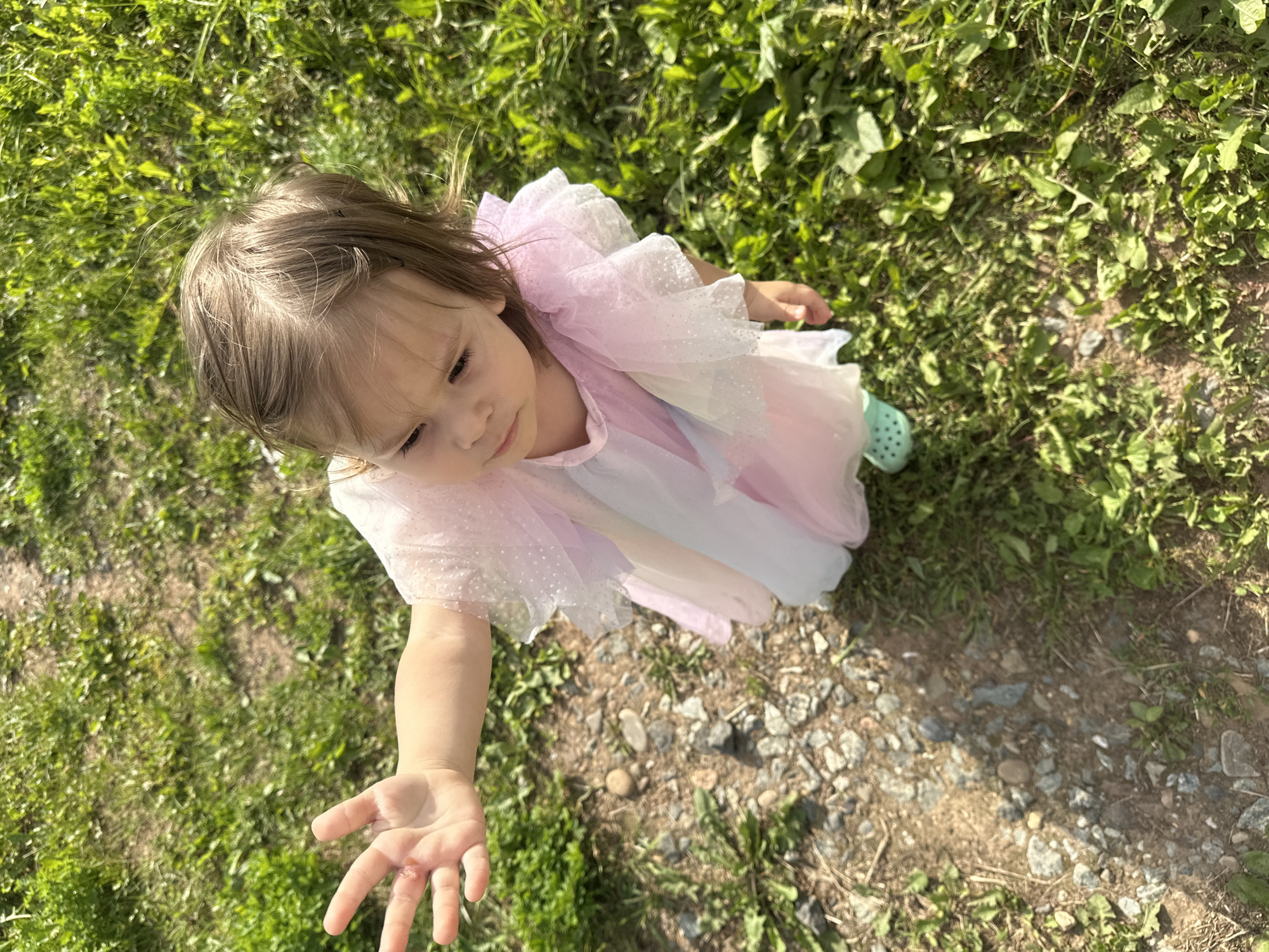 A young child with light brown hair, wearing a pastel-colored dress with sparkly tulle and mint green Crocs, is standing on a path with a gravelly surface and grass on either side. The child's left hand is extended forward and appears to have a minor scrape or abrasion on the palm. The child is looking down and to the right of the frame. The scene is outdoors, with a shallow depth of field, suggesting it was taken in Kharnavy, Russia, on a sunny day.