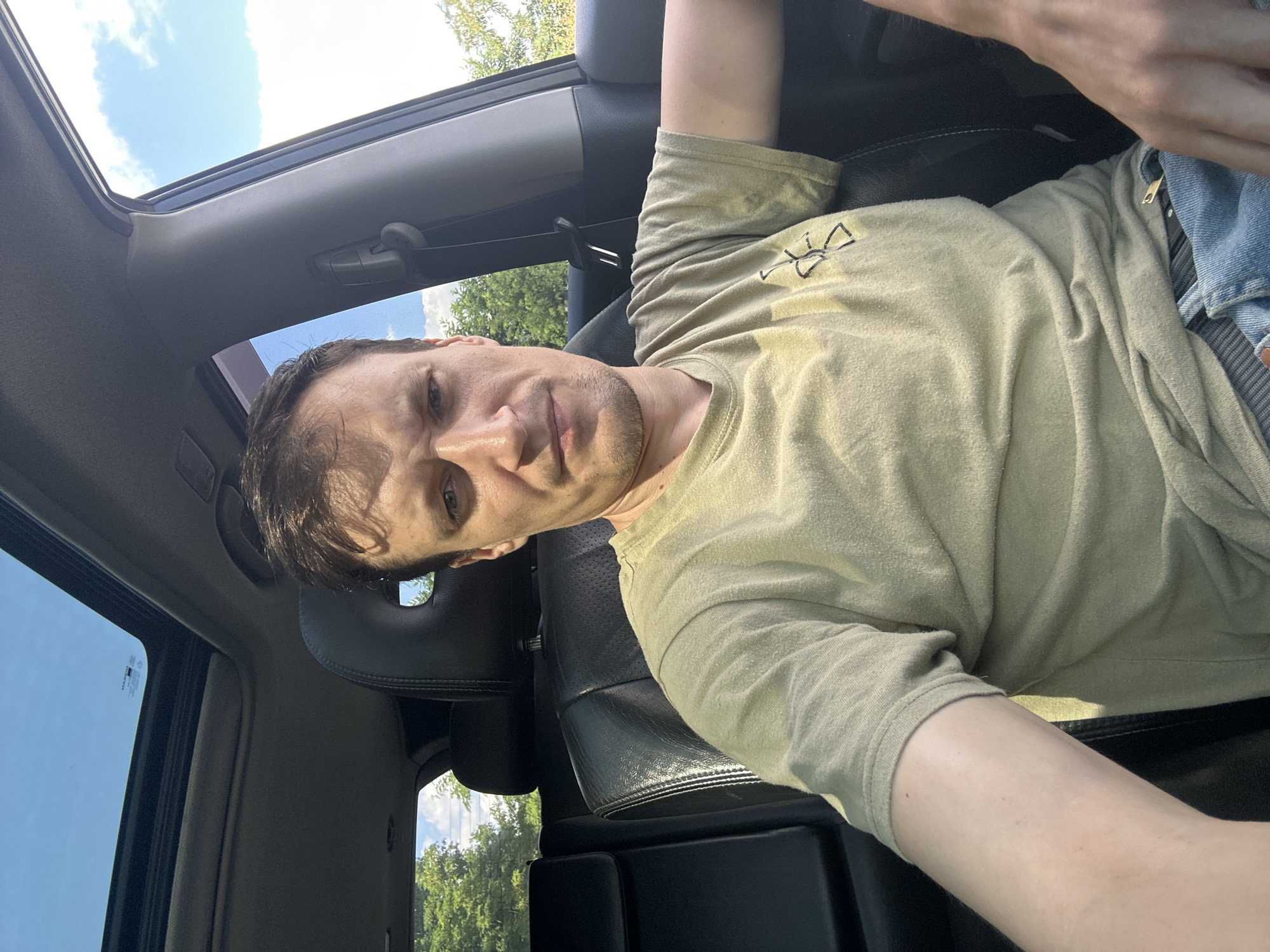 A man is sitting in the driver's seat of a car, looking at the camera. He has short brown hair and is wearing a light green t-shirt. The car has black leather seats and a sunroof. Outside the car, there are trees and a blue sky with some clouds. The scene is lit by natural sunlight. The specific location in the city of Sichyntsi, Ukraine, is not discernible from the image.