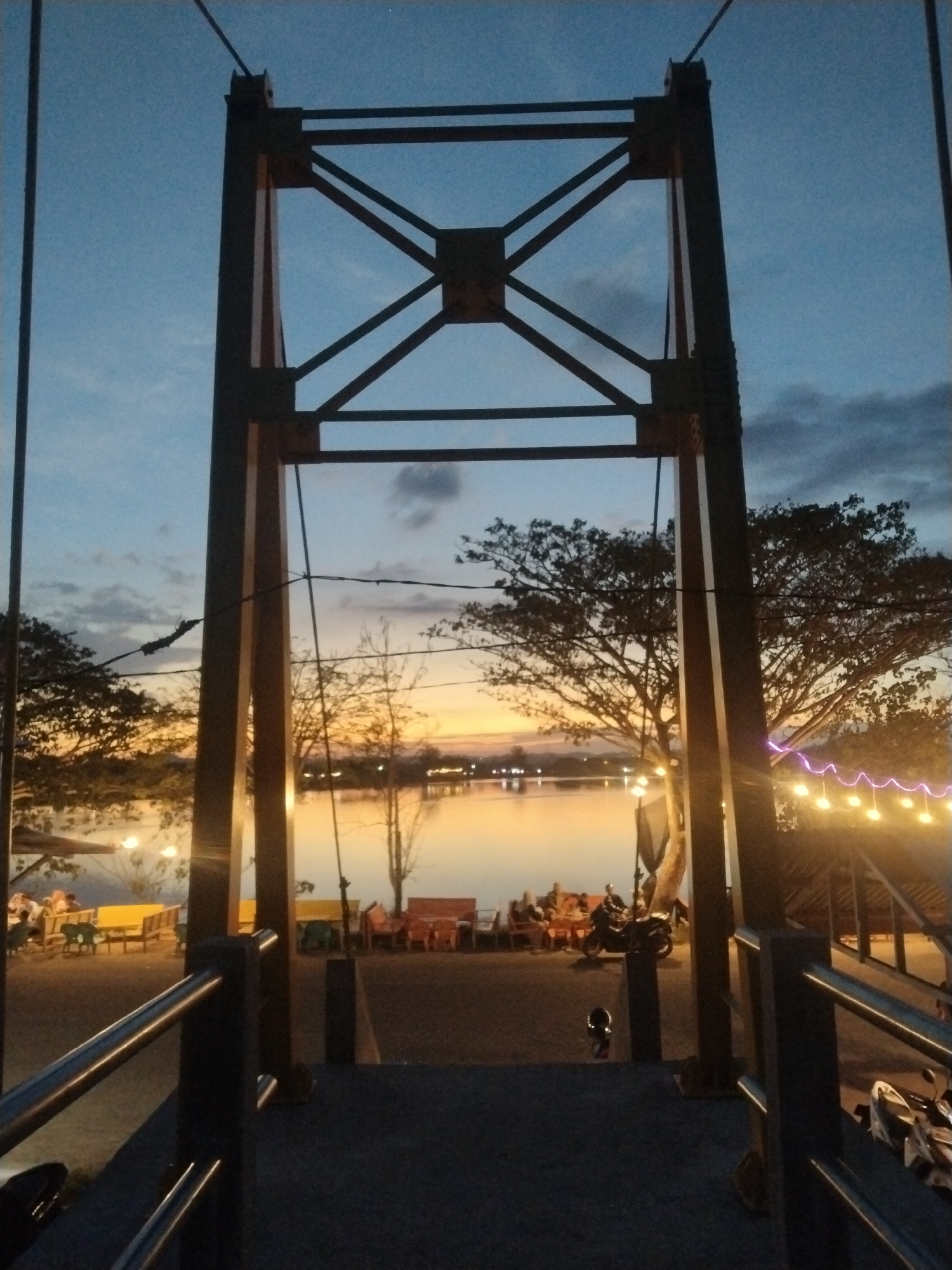 The image depicts a view from a bridge in Lhokseumawe, Indonesia, at twilight. The bridge's metal framework is in the foreground. Through the framework, a body of water reflects the colors of the sunset, with lights visible along the shore. People are seated at tables near the water. A motorcycle is parked nearby. The overall scene is twilight.