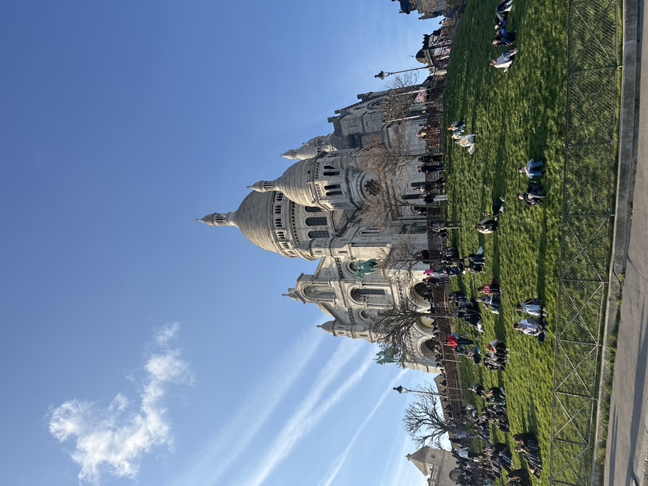 This image displays the Sacré-Cœur Basilica situated on Montmartre, in Paris, France. The large, white-domed basilica occupies the upper portion of the frame, featuring multiple domes, ornate architectural details, and two equestrian statues on its lower facade. The structure rests on a steep, bright green grassy hill, which is populated by numerous individuals. Most people are seated or reclining on the sunlit grass, facing generally away from the viewer, while others are standing closer to the basilica's base. They appear to be casually dressed and engaged in leisure. A low, dark green metal fence runs horizontally across the lower foreground, separating the grassy slope from a concrete pathway. Bare deciduous trees are visible on the hillside near the basilica. The sky is clear blue with sparse, wispy clouds and several distinct parallel contrails.