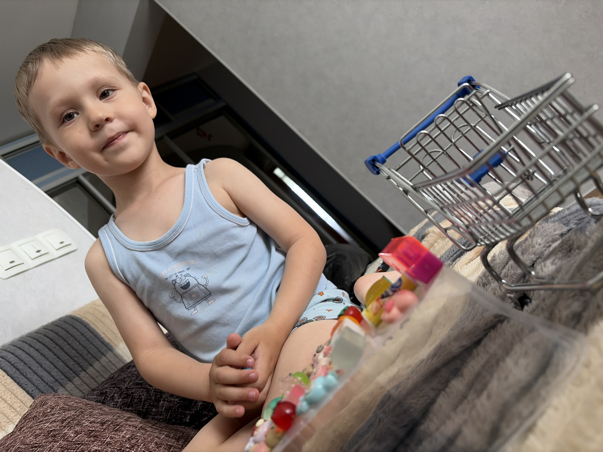 A young boy is captured in an indoor setting, likely a home in Chaikovsky, Russia. The main subject, a toddler or preschooler with light brown hair, looks directly at the camera with a slight, gentle smile. He is wearing a light blue sleeveless top adorned with a cartoon robot graphic and the partially visible text "I'M LITTLE ROBOT-MAN". His attire also includes light blue patterned shorts.
He is seated on a multi-toned, striped cushion or blanket with shades of brown, grey, and beige. His hands are clasped together in front of him. To the right of the frame, a miniature shopping cart, metallic silver with blue plastic accents, rests on a fluffy grey blanket. Adjacent to the boy's leg and near the shopping cart is a clear plastic container or bag filled with various colorful items, appearing to be candies or small toys like gummies and beads.
The background features white light switches on a light-colored wall to the left, and a dark doorway or furniture item further back. The ceiling is a plain light grey. The scene conveys a relaxed, playful atmosphere, suggesting a moment of leisure or play at home. The indoor lighting does not indicate a specific time of day.
