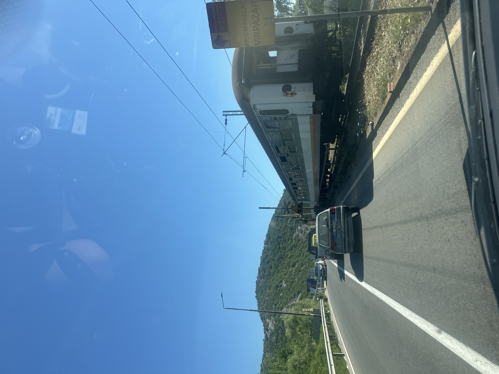 The image shows a scene near Virpazar, Montenegro, featuring a train adjacent to a road. A train carriage is visible, with a sign reading "Pruzni Prelaz Virpazar" (Railway Crossing Virpazar). Several cars are on the road, with one car's license plate, "BR AS249," partially visible. The background includes a hillside covered in greenery and a clear blue sky. The photo appears to have been taken from inside a vehicle, as evidenced by reflections on the window. Overhead power lines for the train are present.