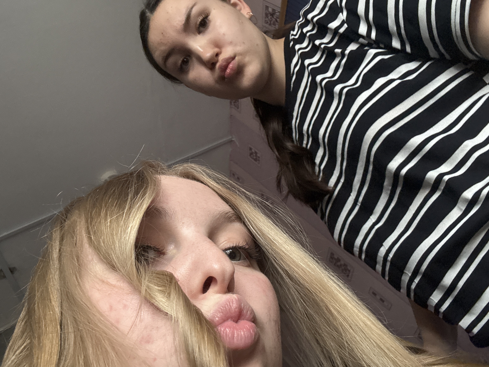 Two young women pose for a selfie. One has long blonde hair that partially obscures her face, and she is puckering her lips. The other woman has darker hair pulled back into a braid and is wearing a black and white striped shirt. She also is puckering her lips. The background is indistinct but appears to be an interior setting. There are no visible elements to definitively place the scene in the city of Perm, Russia.
